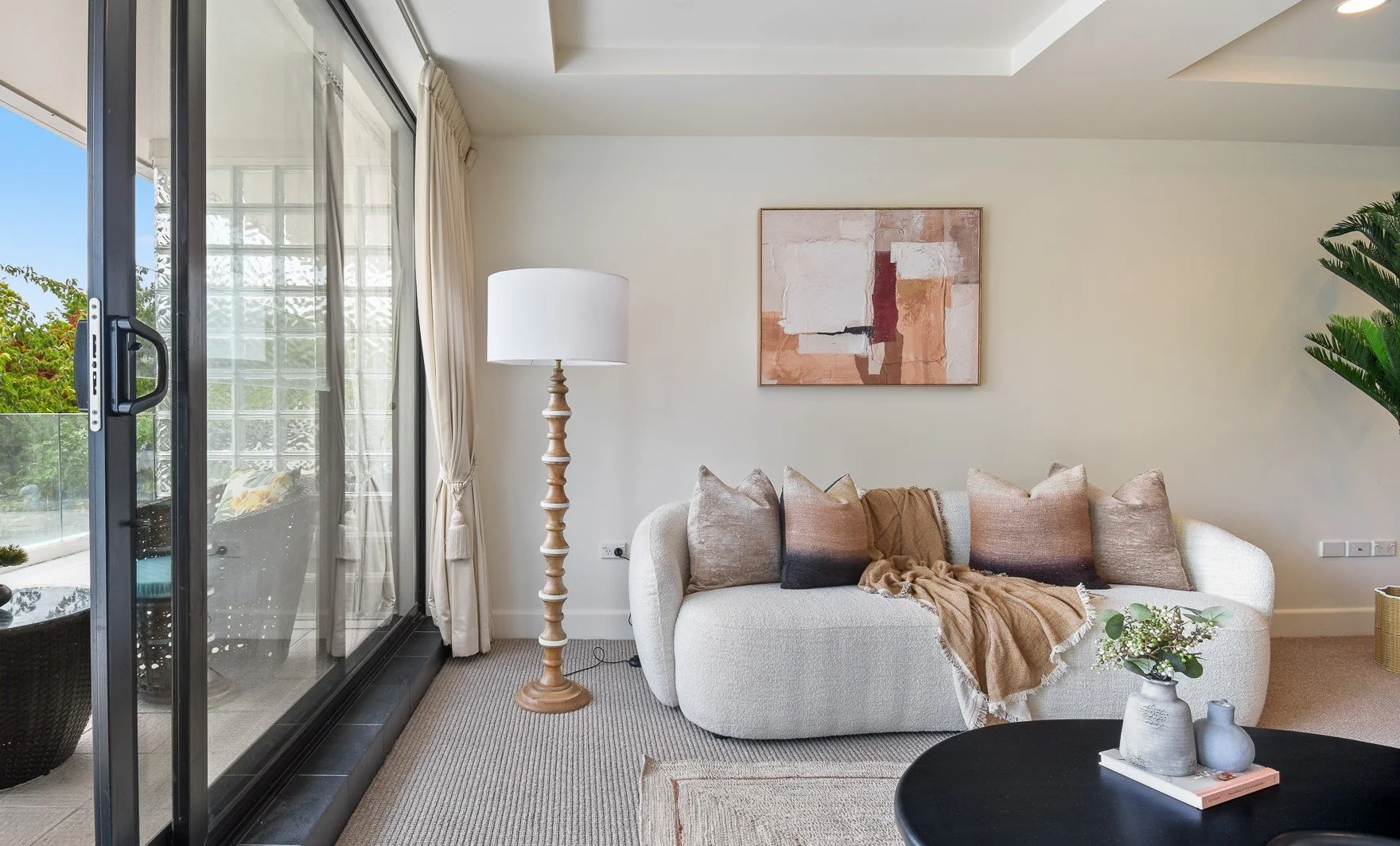 Living room with light-colored sofa, beige throw blanket, five decorative pillows, abstract artwork, tall wooden floor lamp, sliding glass door leading to balcony with outdoor seating and greenery, small table with vases and book, beige carpet, cream