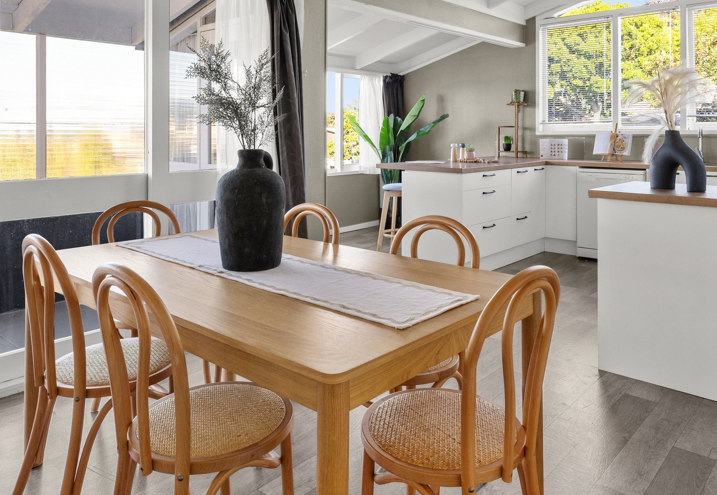 A bright dining area with a wooden table, six matching chairs, and a large black vase with dried branches as a centerpiece. The room features large windows with a view of green trees, white kitchen cabinets with wooden countertops, and decorative vas