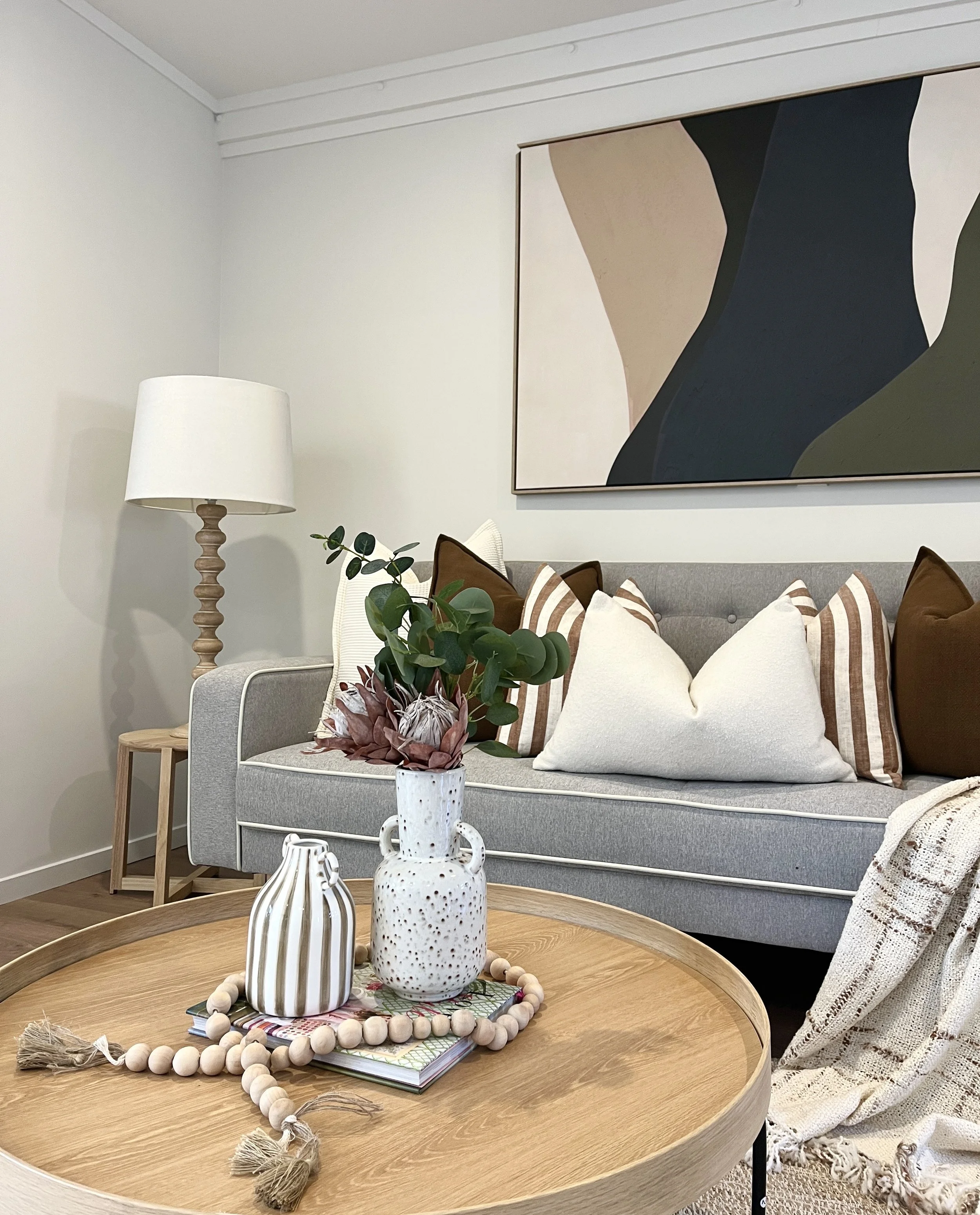 Living room with light gray sofa, brown and striped pillows, large abstract wall art, wooden side table with a lamp, wooden coffee table with decorative vases, plant, and tray with beads, and a blanket draped over an armchair.