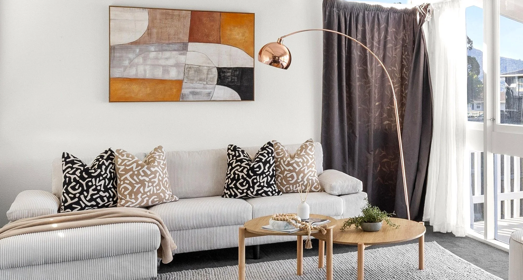 Living room with white striped sofa, decorative pillows, a wooden coffee table with decor, a tall copper arc floor lamp, abstract wall art, dark curtains, and sliding glass door with an outdoor balcony view.