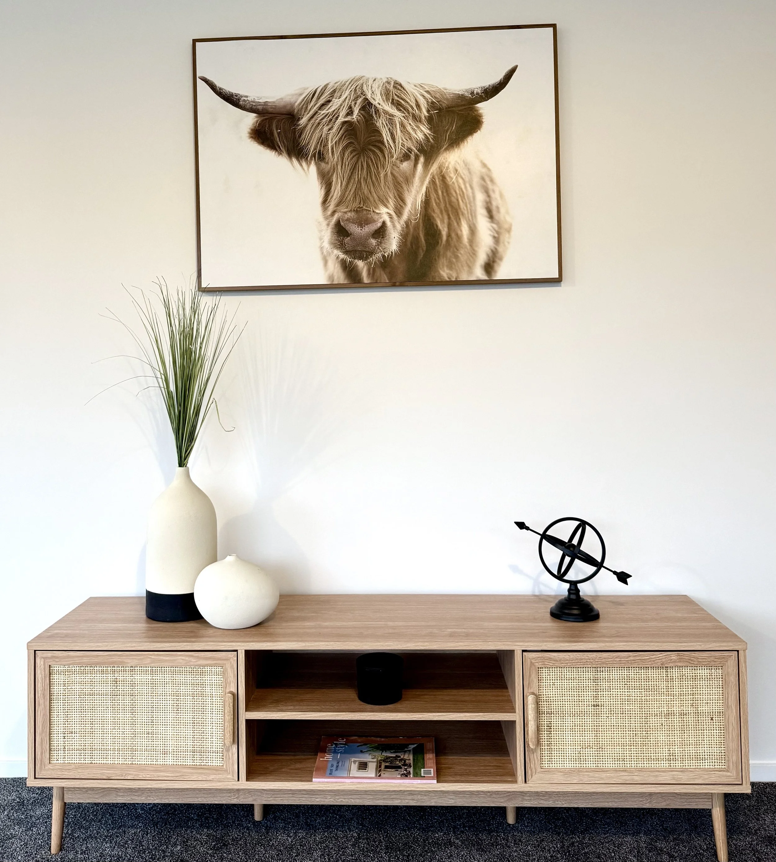 Living room with a wooden console table, a white vase with tall green grass, a smaller white round vase, a decorative armillary sphere, a framed cow photograph on the wall, and a magazine on the shelf.