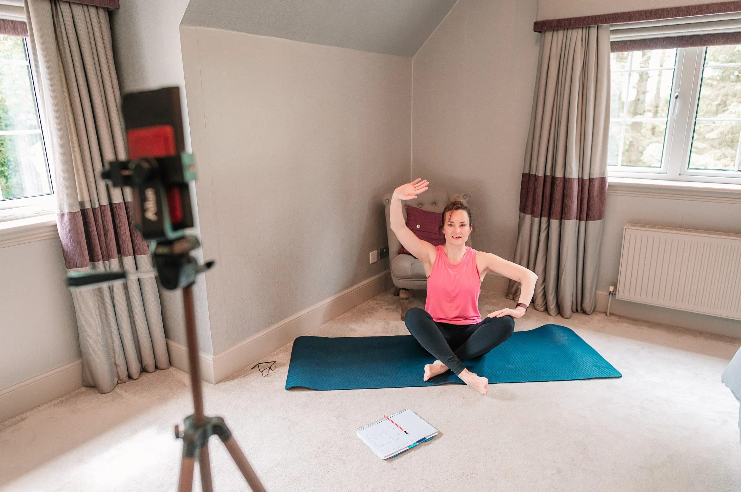 A woman in a pink athletic top and black leggings sitting on a blue yoga mat in a room with beige carpet, beige walls, and large windows with curtains, recording a hypopressives video with a camera on a tripod.