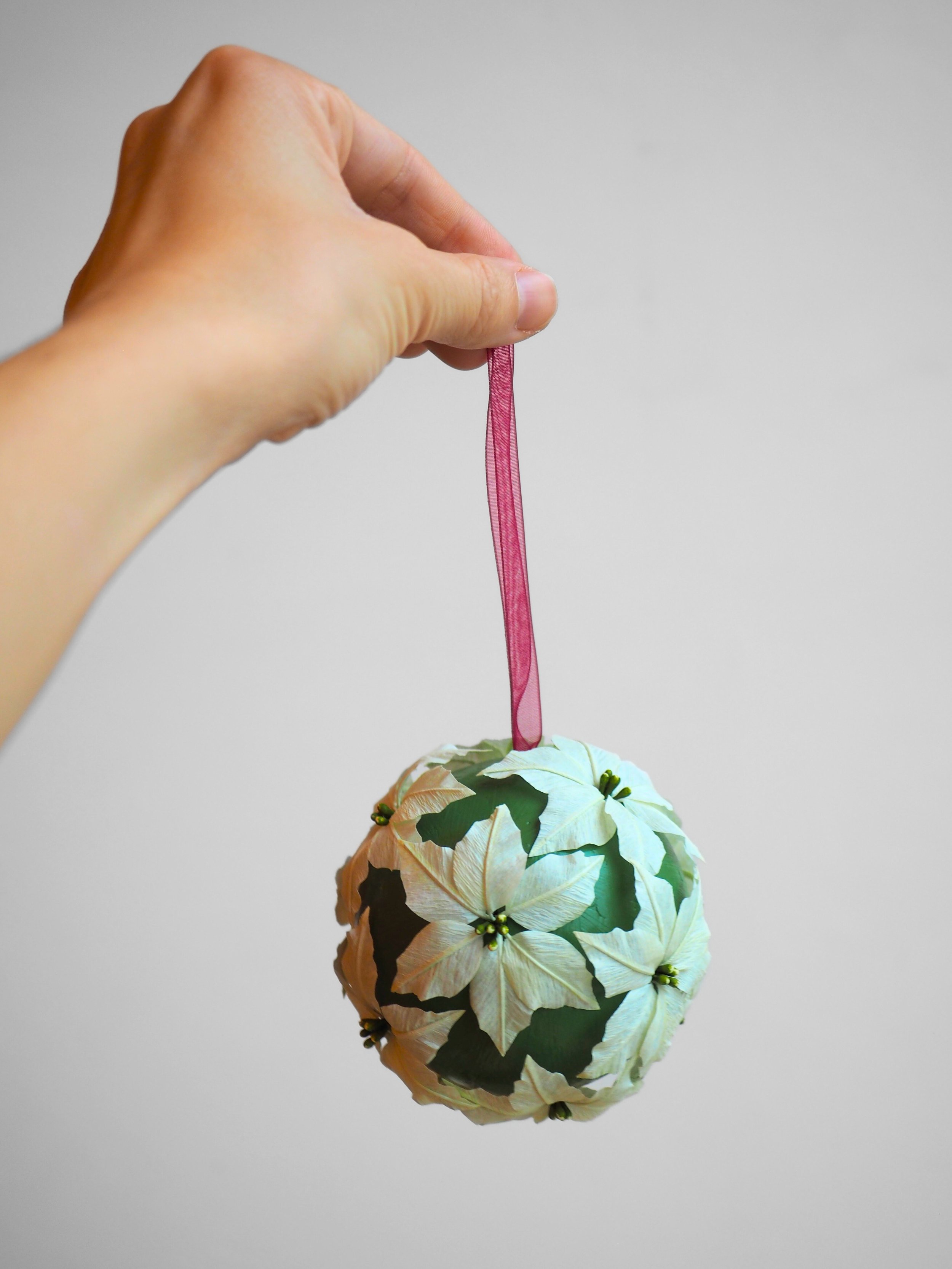 A hand holding a pink ribbon attached to a decorative spherical ornament with a poinsettia flower design.