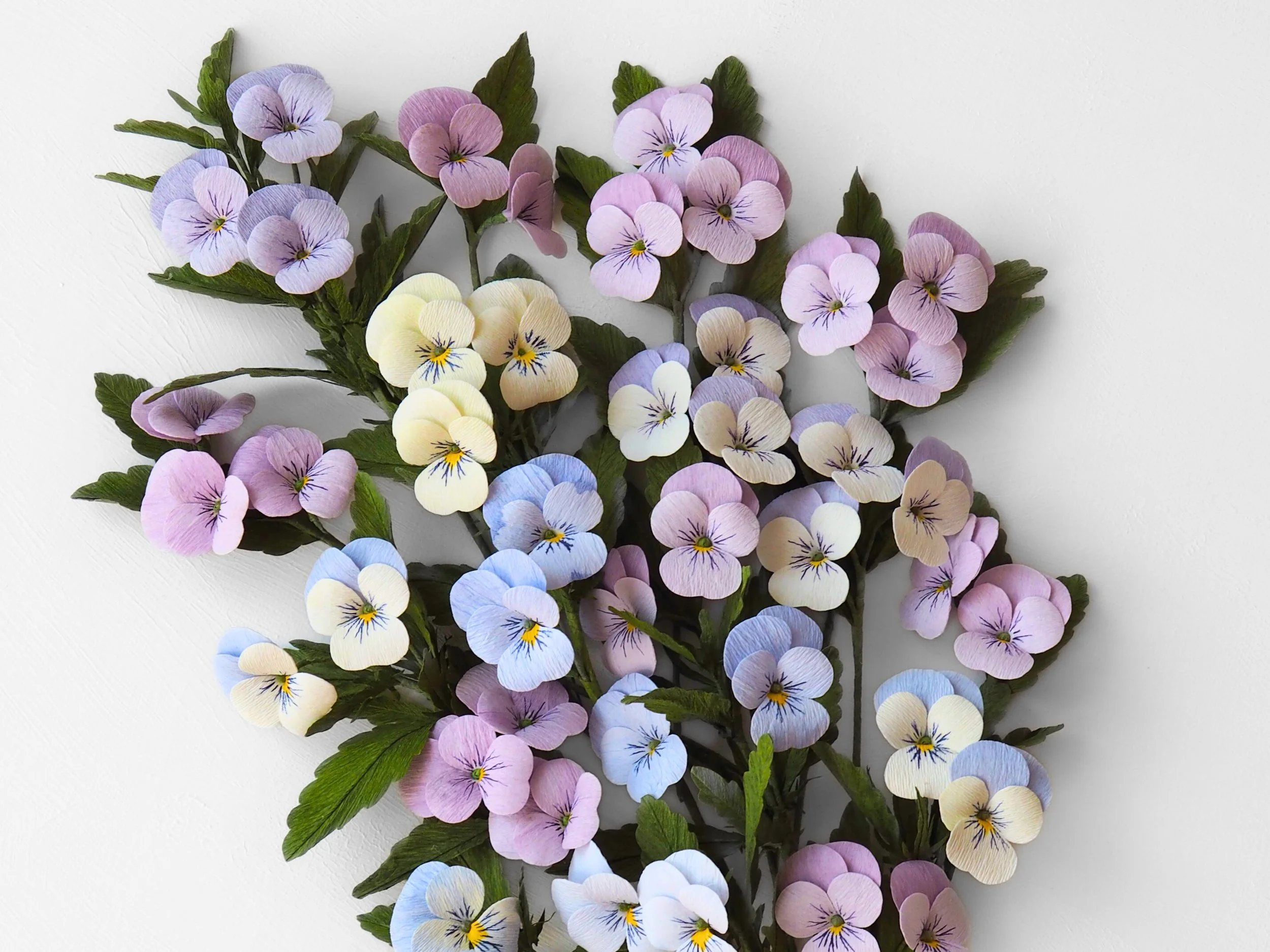 Arrangement of colorful paper pansy flowers in pastel shades of purple, yellow, blue, and white with green leaves against a white background.