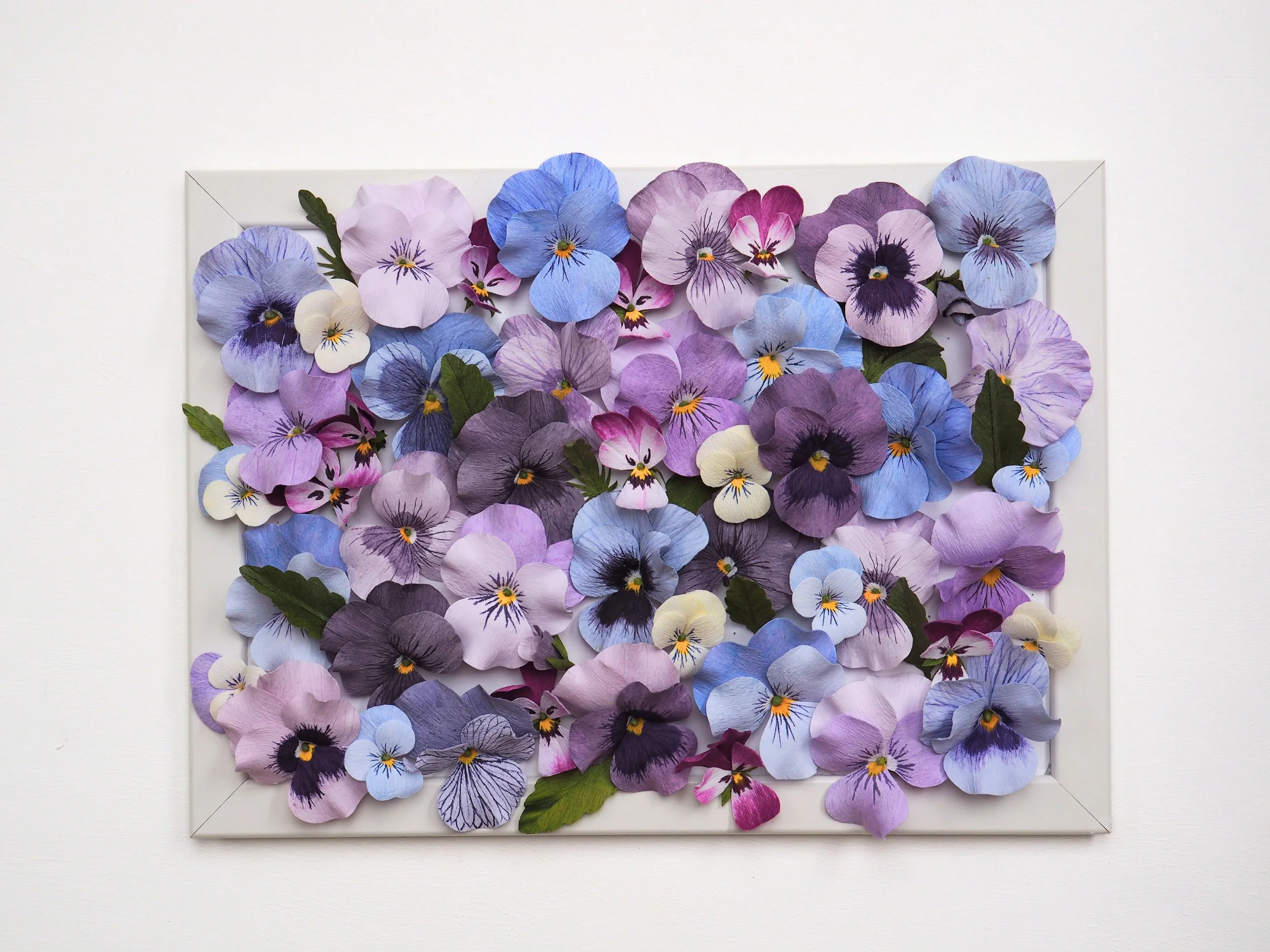A framed collection of artificial flowers in shades of purple, blue, pink, cream, and green leaves arranged on a white background.