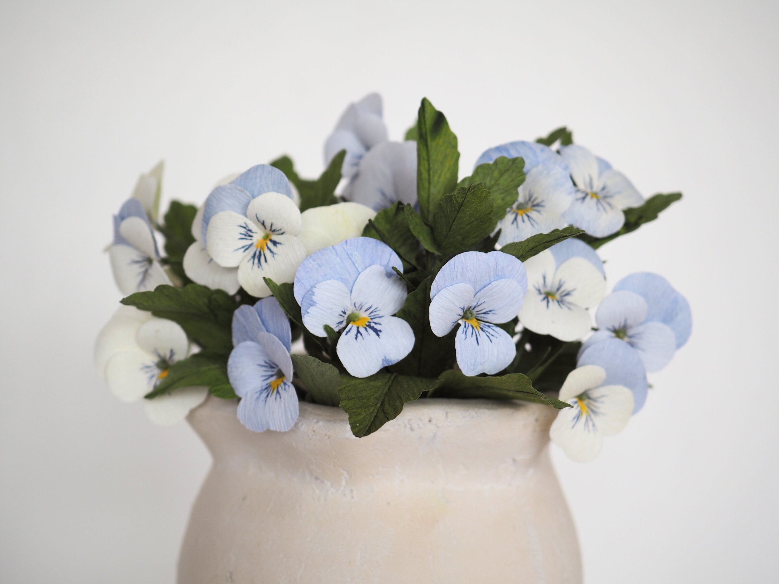 A white ceramic vase filled with light blue and white artificial pansy flowers with green leaves.