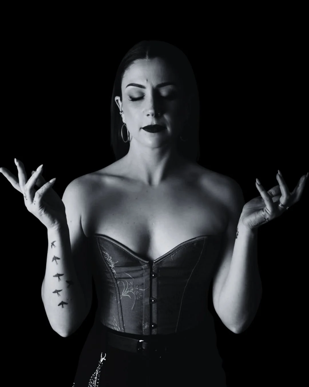 A woman with dark hair and makeup, wearing a strapless corset and hoop earrings, has her eyes closed and hands raised with fingers spread, against a black background.