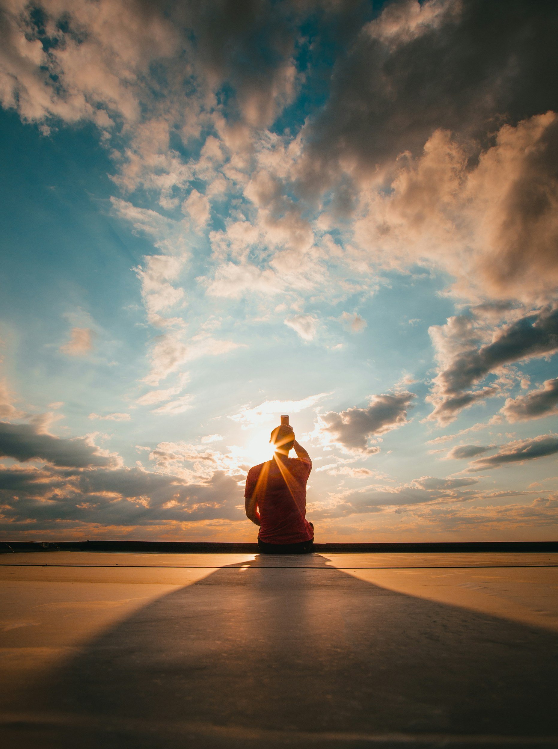 Person sitting on a dock taking a photo of the sunset sky with clouds and the sun partially obscured behind their head.