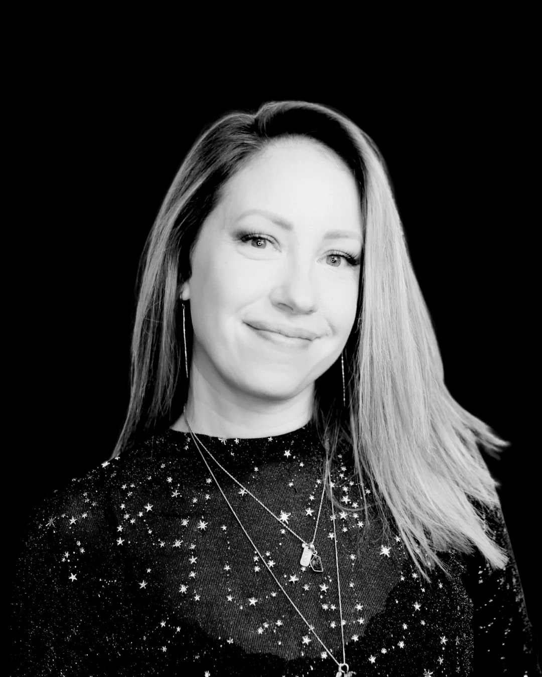 Black and white portrait of a woman with long hair, smiling, wearing star-patterned clothing and layered necklaces against a black background.