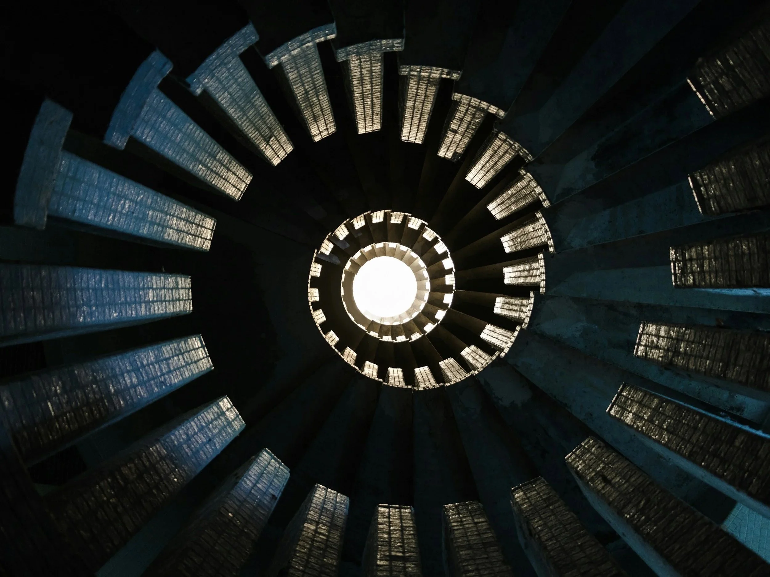 Looking up through the center of a circular staircase with metal steps, with light shining from the top.