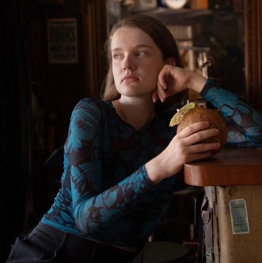 A young woman with long brown hair, wearing a blue patterned shirt, rests her chin on her hand while sitting at a wooden bar or table. She holds a coconut drink with a small umbrella in her right hand. She looks contemplatively to her left in a dimly lit setting with bookshelves in the background.