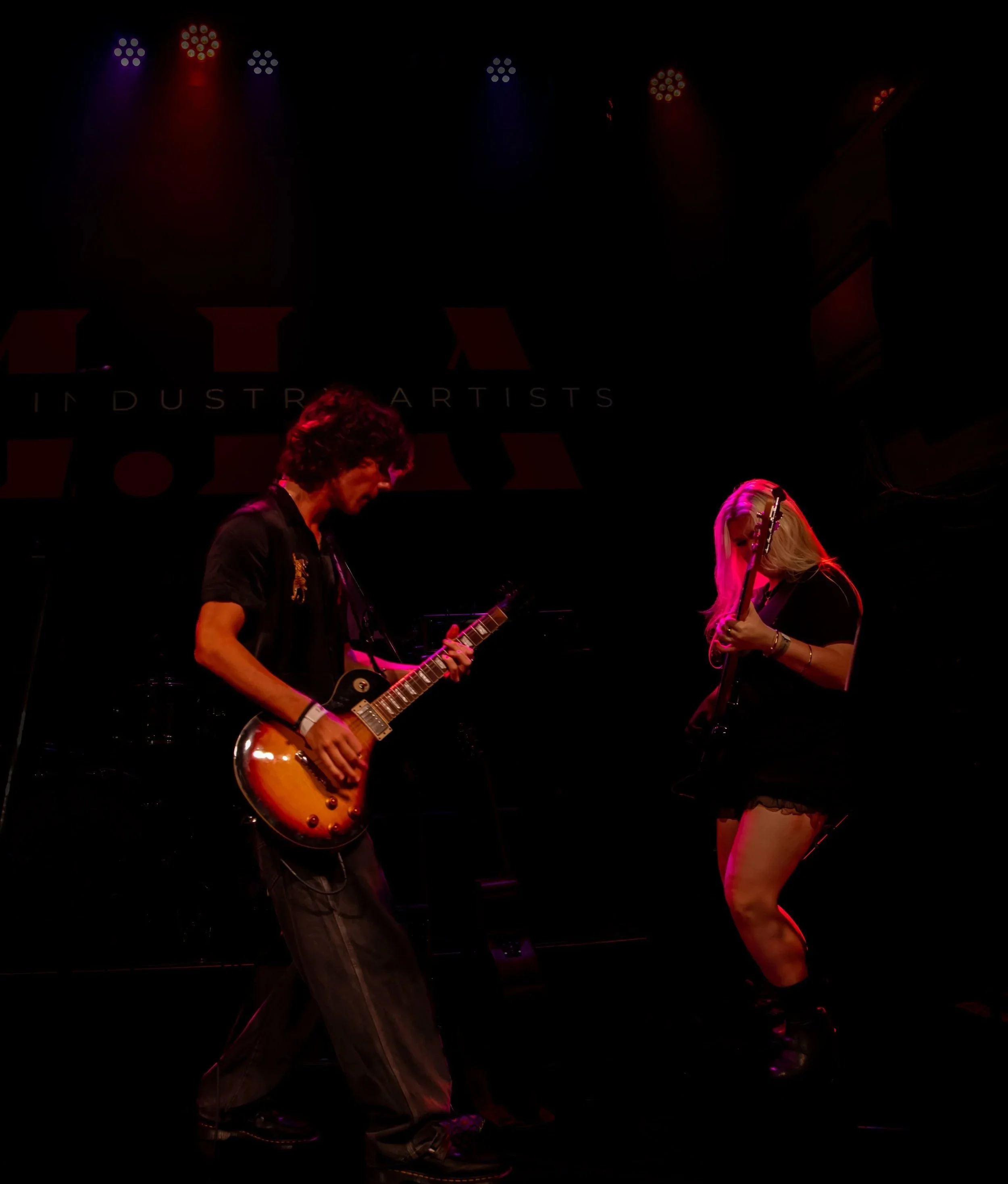 A musician playing an electric guitar on stage, with stage lighting above and a dark background.