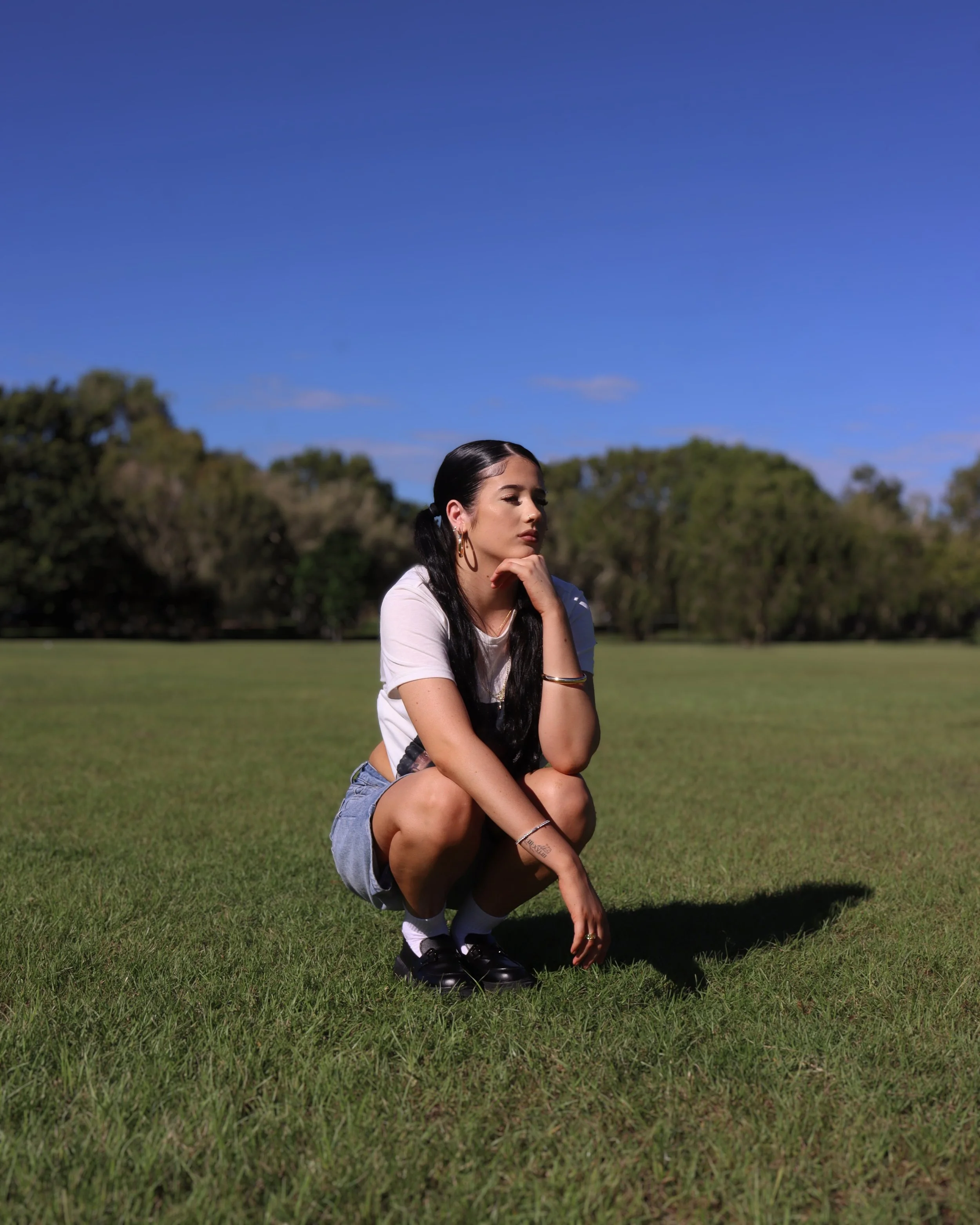 A young woman crouching on the grass with trees in the background and a blue sky above.