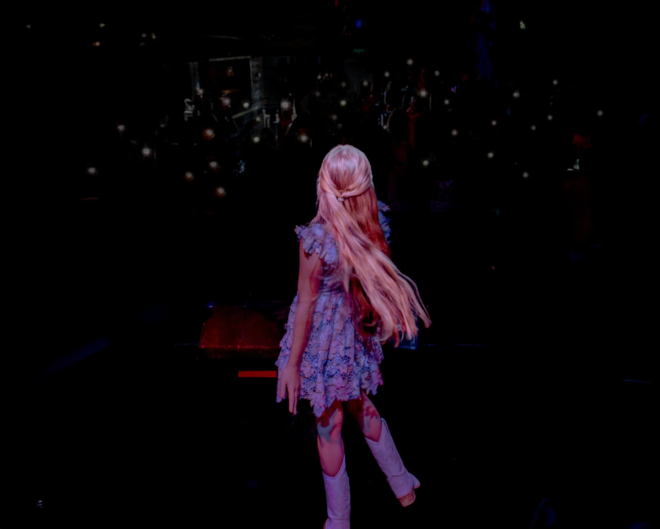 A person with long, pink hair wearing a floral dress and white boots standing on stage in front of a dark background with small, bright lights.