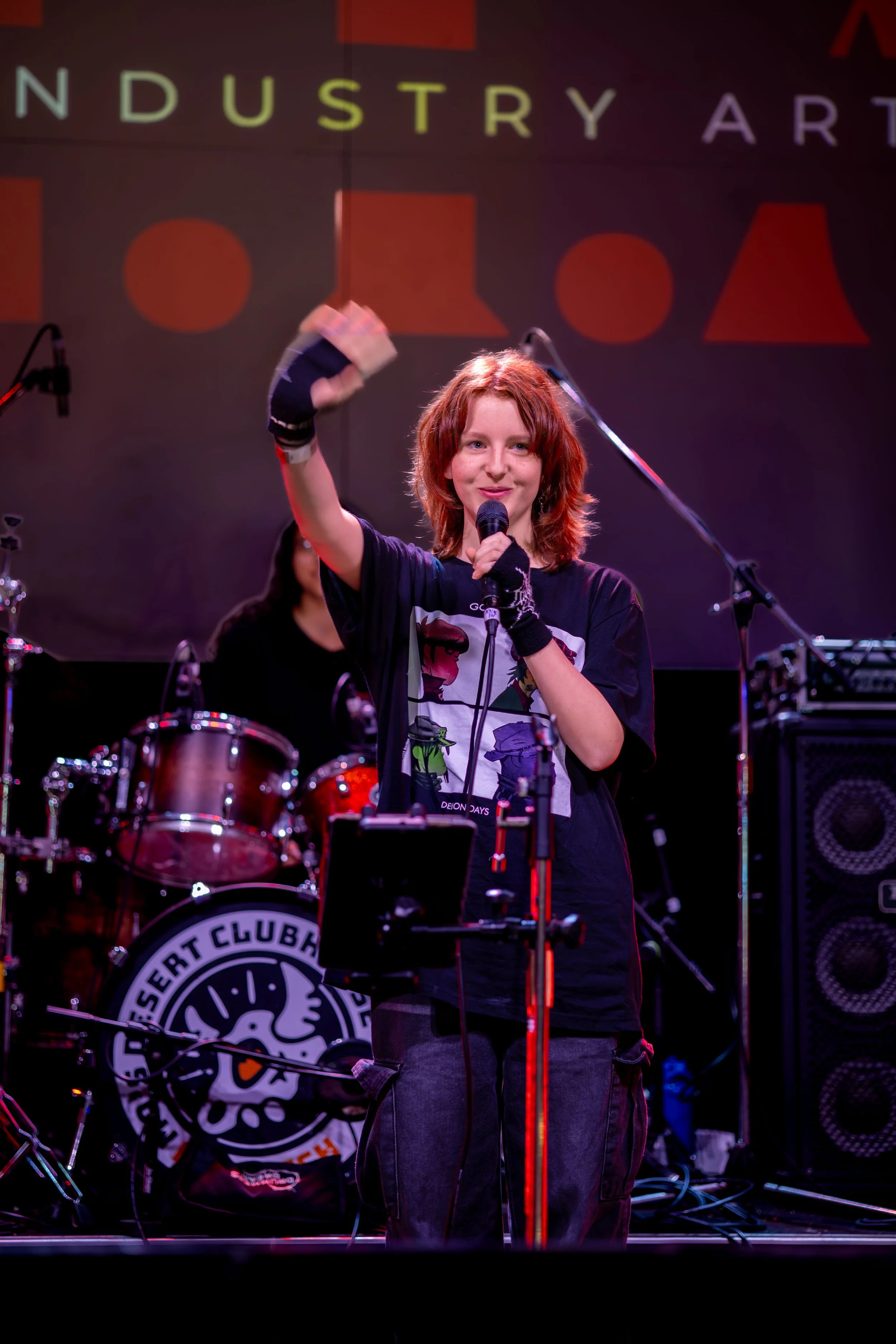 A young person with red curly hair singing into a microphone on a stage, with a drum set and microphone stands behind them, and a background displaying the text 'INDUSTRY ART'.