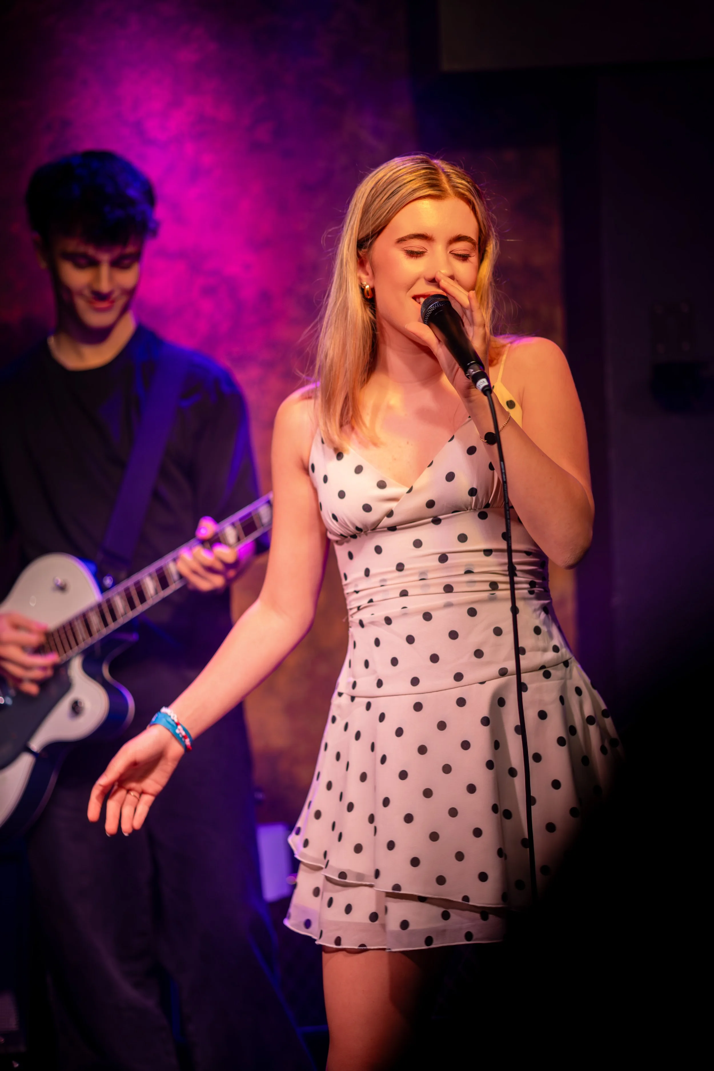 A young woman singing into a microphone on stage, wearing a polka dot dress and a young man playing guitar in the background.