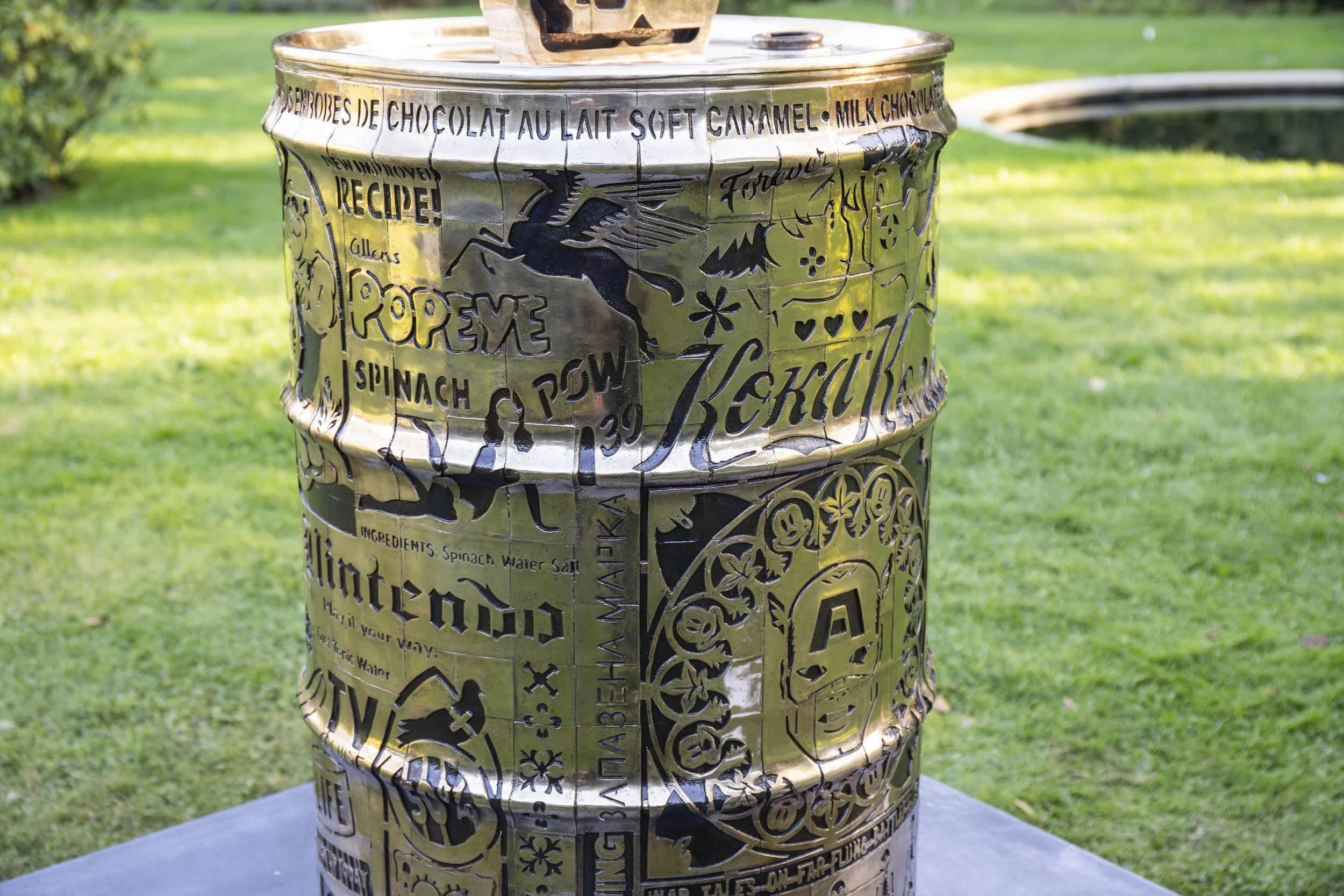 Close-up of the bronze oil drum base of Lhouette's Angel Cake sculpture, showing intricate stencil-derived engravings, lettering and layered graphic imagery.