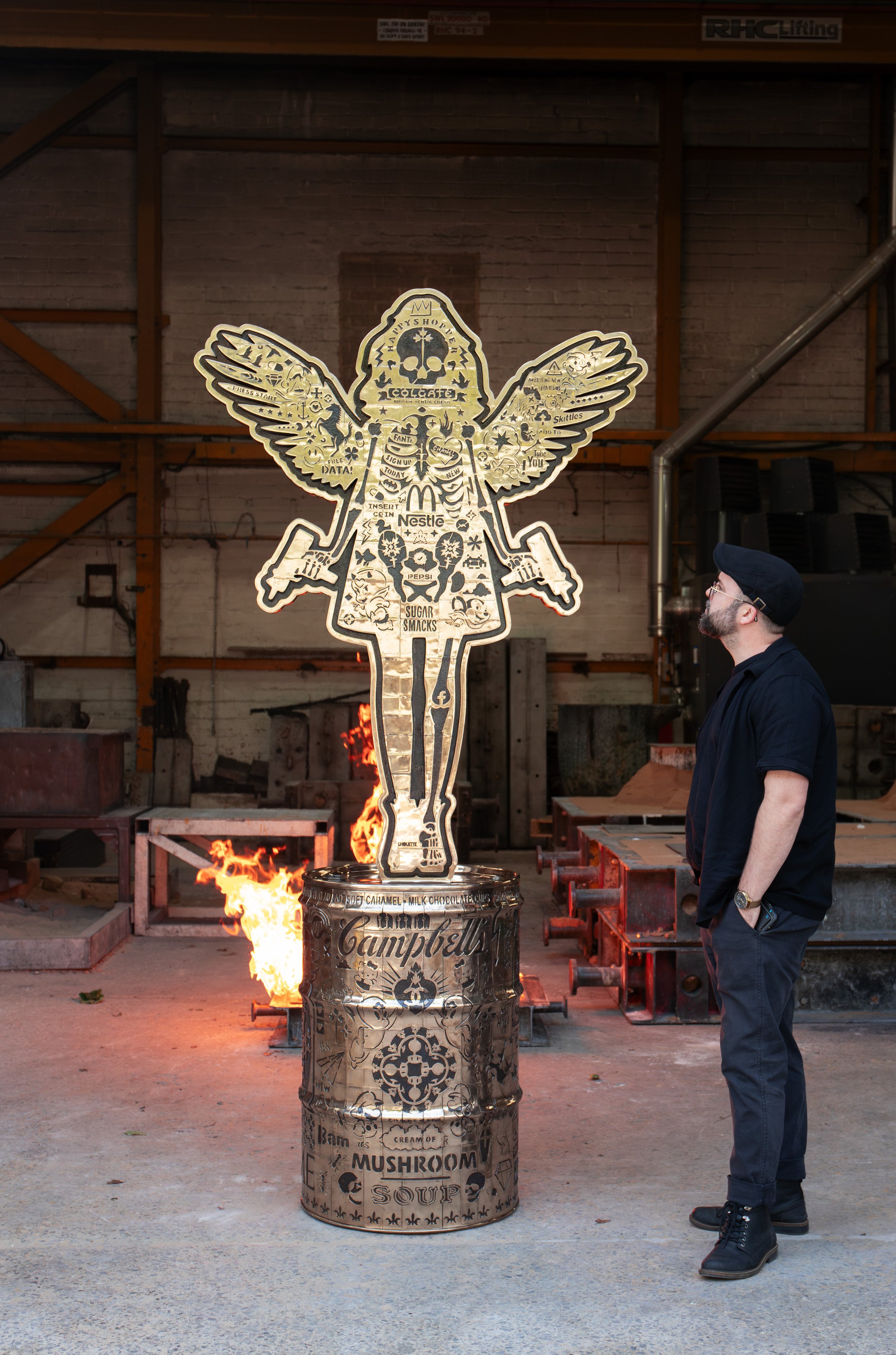 Contemporary artist Lhouette standing beside his completed Angel Cake bronze sculpture at Pangolin Editions foundry, with the full-scale winged figure mounted on a cast bronze oil drum.
