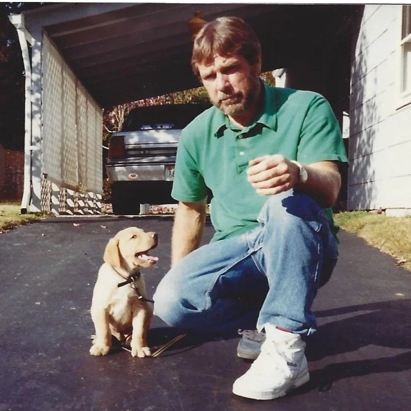 Eddie Busi inspiration behind Oaked Interiors interior design studio, crouching next to a yellow Labrador puppy (Duke) on a driveway, with a truck and house in the background.