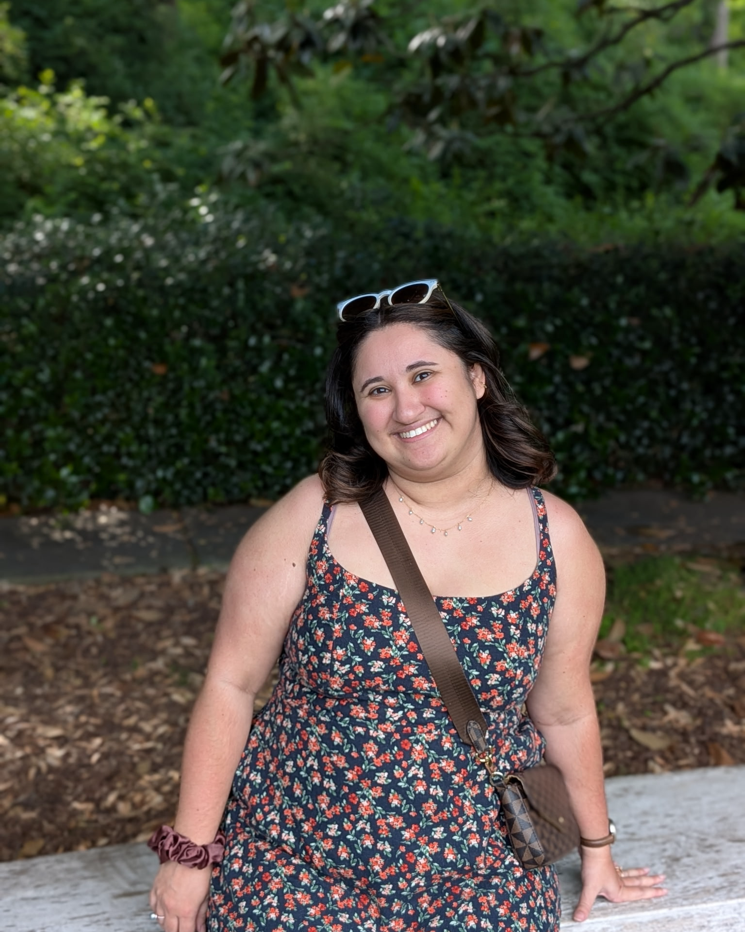 Sara Masoudi-Grasso of Oaked Interiors, interior design,  with dark hair, wearing sunglasses on her head, a floral dress, and a brown crossbody bag, sitting outdoors on a wooden bench or ledge, smiling with a background of green trees and bushes.