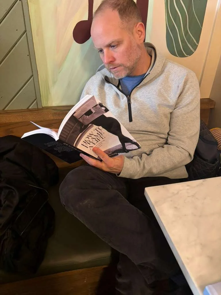 A man with short hair and a beard sitting in a cafe, reading a magazine. He's wearing a light gray zip-up jacket and dark pants, with a black backpack beside him. The background includes a wall with artistic designs and a small table in front of him.