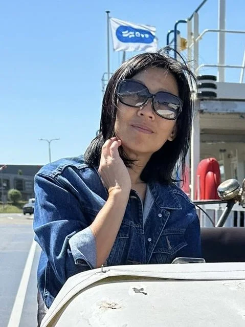 Woman with short black hair wearing sunglasses and a denim jacket, sitting outdoors with a bright blue sky and a flag in the background.