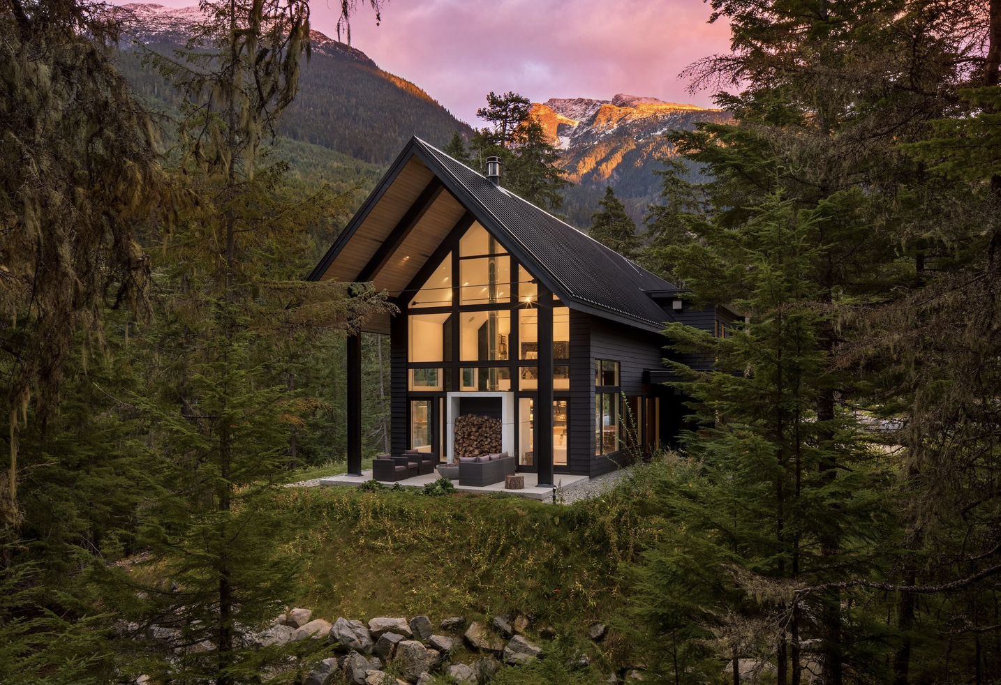 Modern black house with large glass windows and a fireplace with stacked logs, situated among tall green trees with mountain peaks and a pink sky in the background.