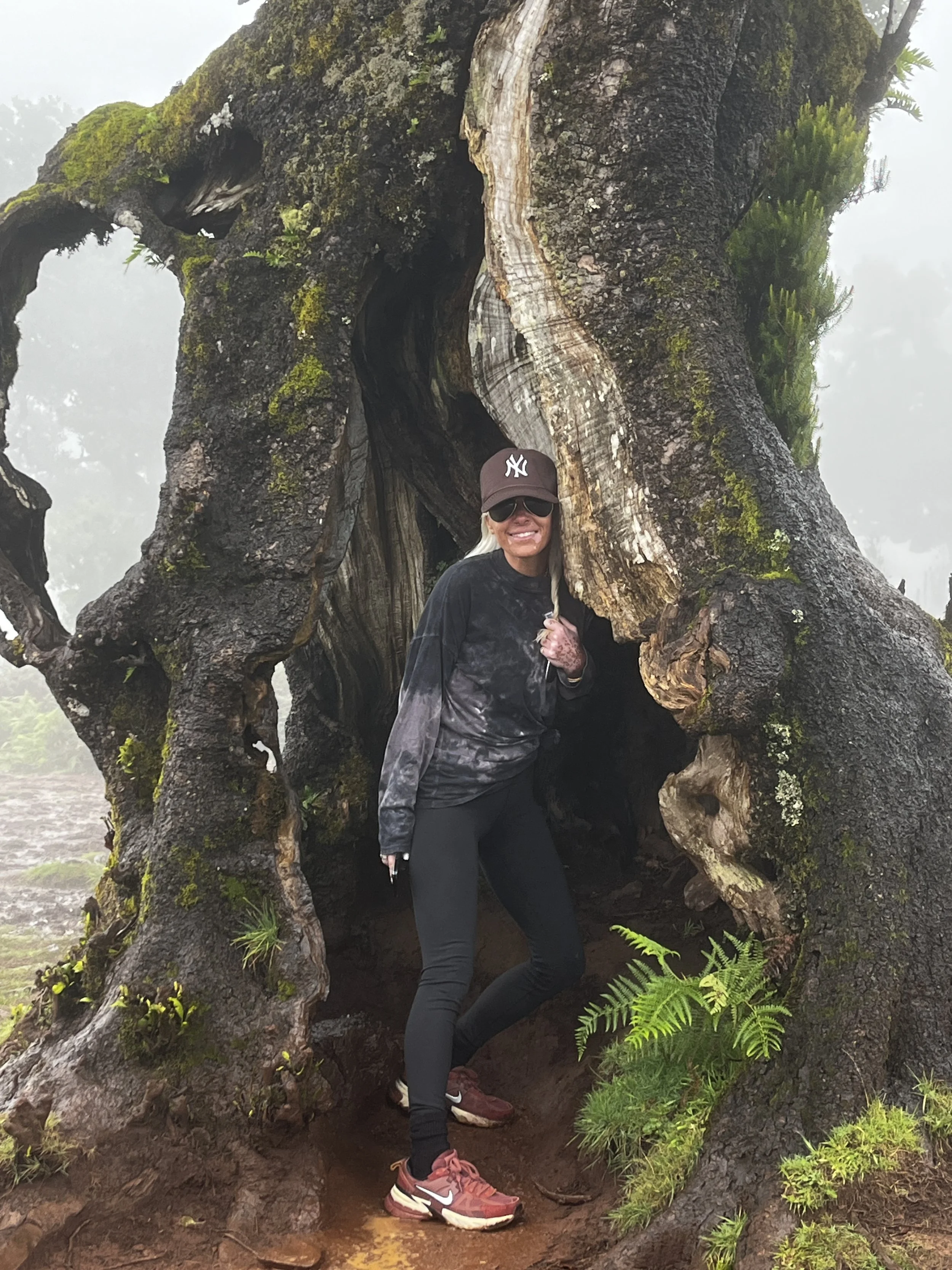 Woman posing next to a large, hollow, moss-covered tree trunk in a foggy outdoor environment, wearing sunglasses, a cap, and hiking attire.
