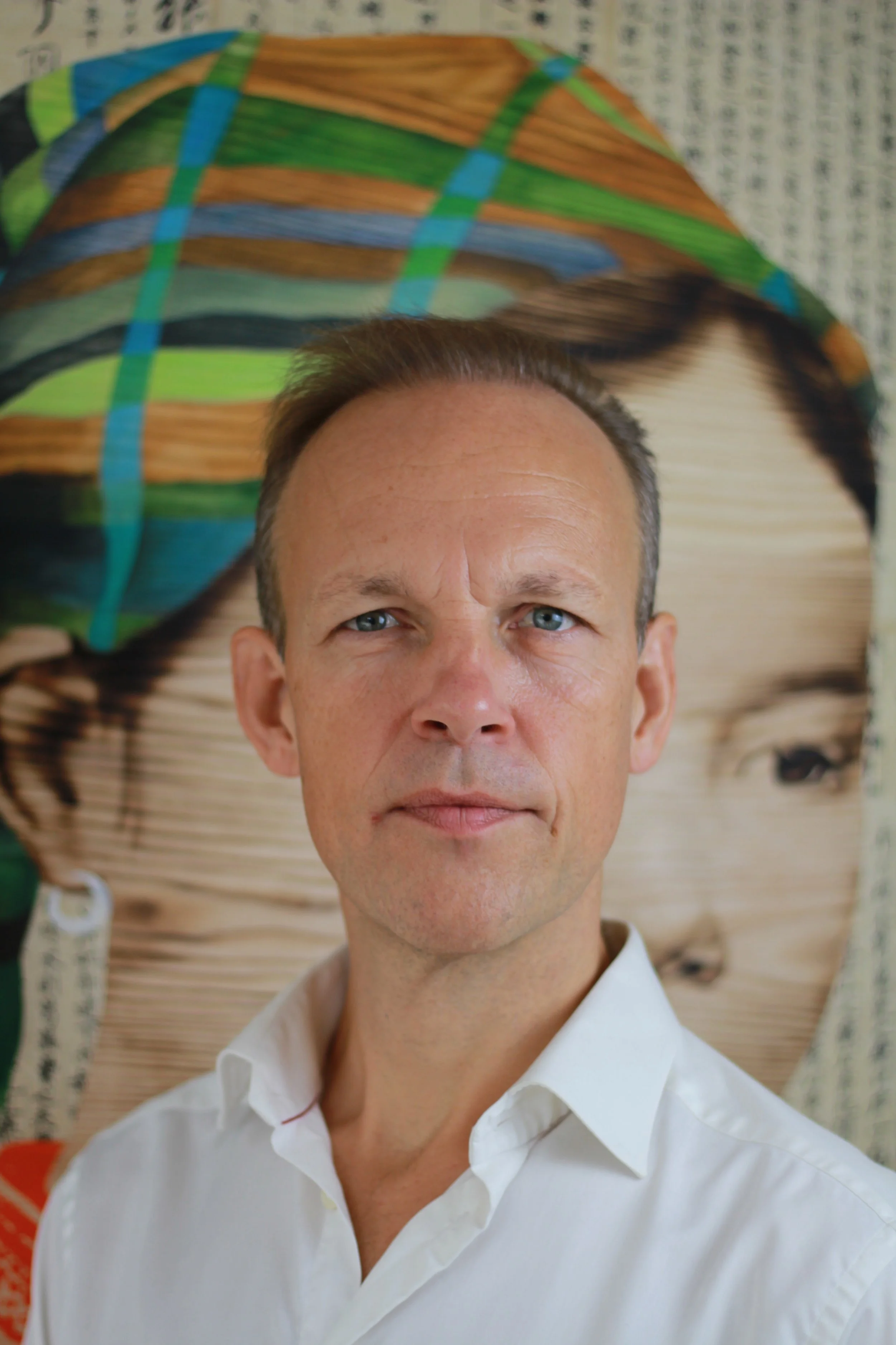 A man with short brown hair and blue eyes wearing a white collared shirt, standing in front of a colorful artwork of a woman with a patterned headscarf.