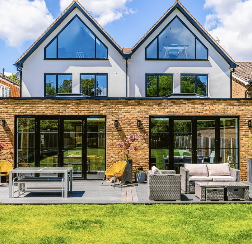 Modern house with white upper floors, brick lower floor, large black-framed glass doors and windows, outdoor patio with yellow chairs, gray sofa and table, small tree with pink blossoms, green lawn, under a blue sky with clouds.