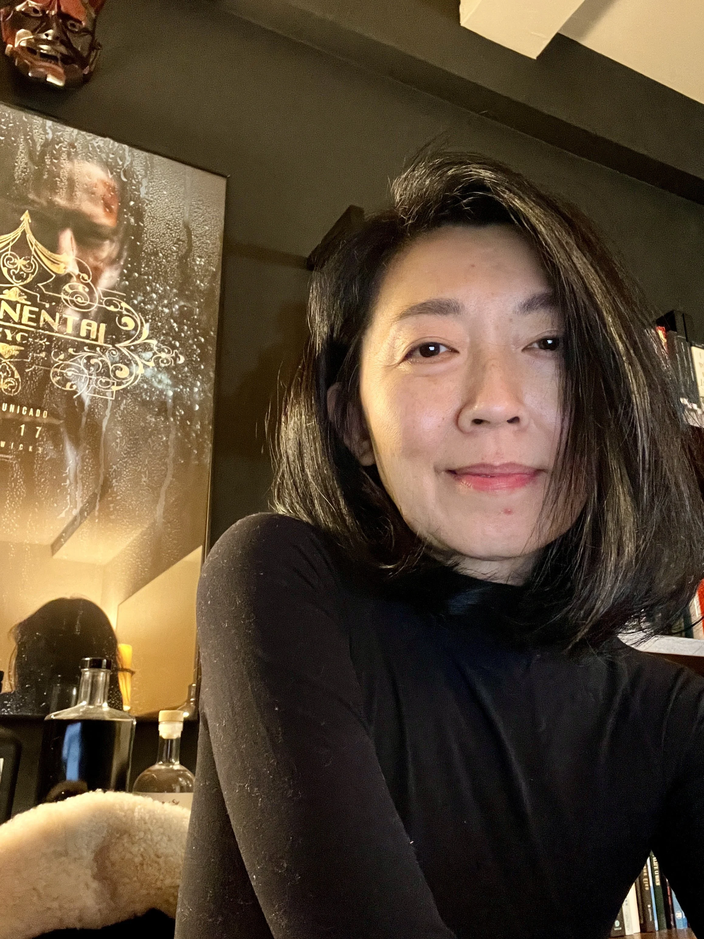 A woman with shoulder-length black hair and a black long-sleeve shirt sitting indoors. Behind her, a poster with gold and white details and some bottles on a table are visible.