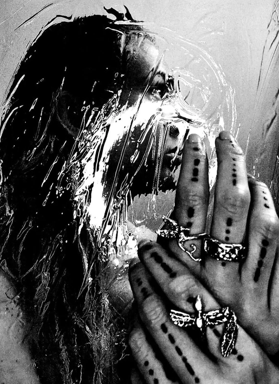 A person with tattoos and rings in front of a window with water on it, touching their face with fingers, looking thoughtfully.