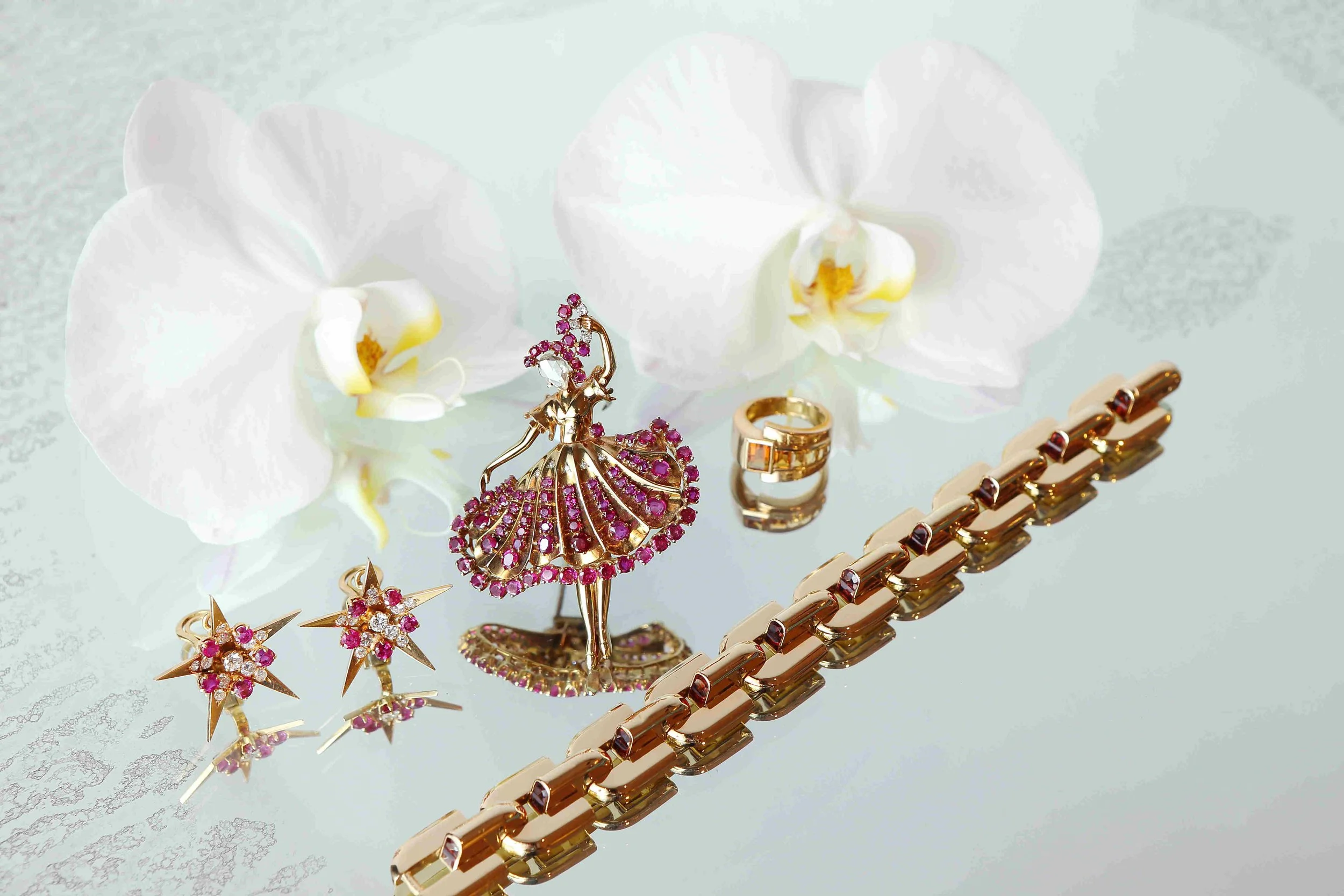 Jewelry set with a ballerina brooch, earrings, ring, bracelet, and white orchids on a reflective surface.
