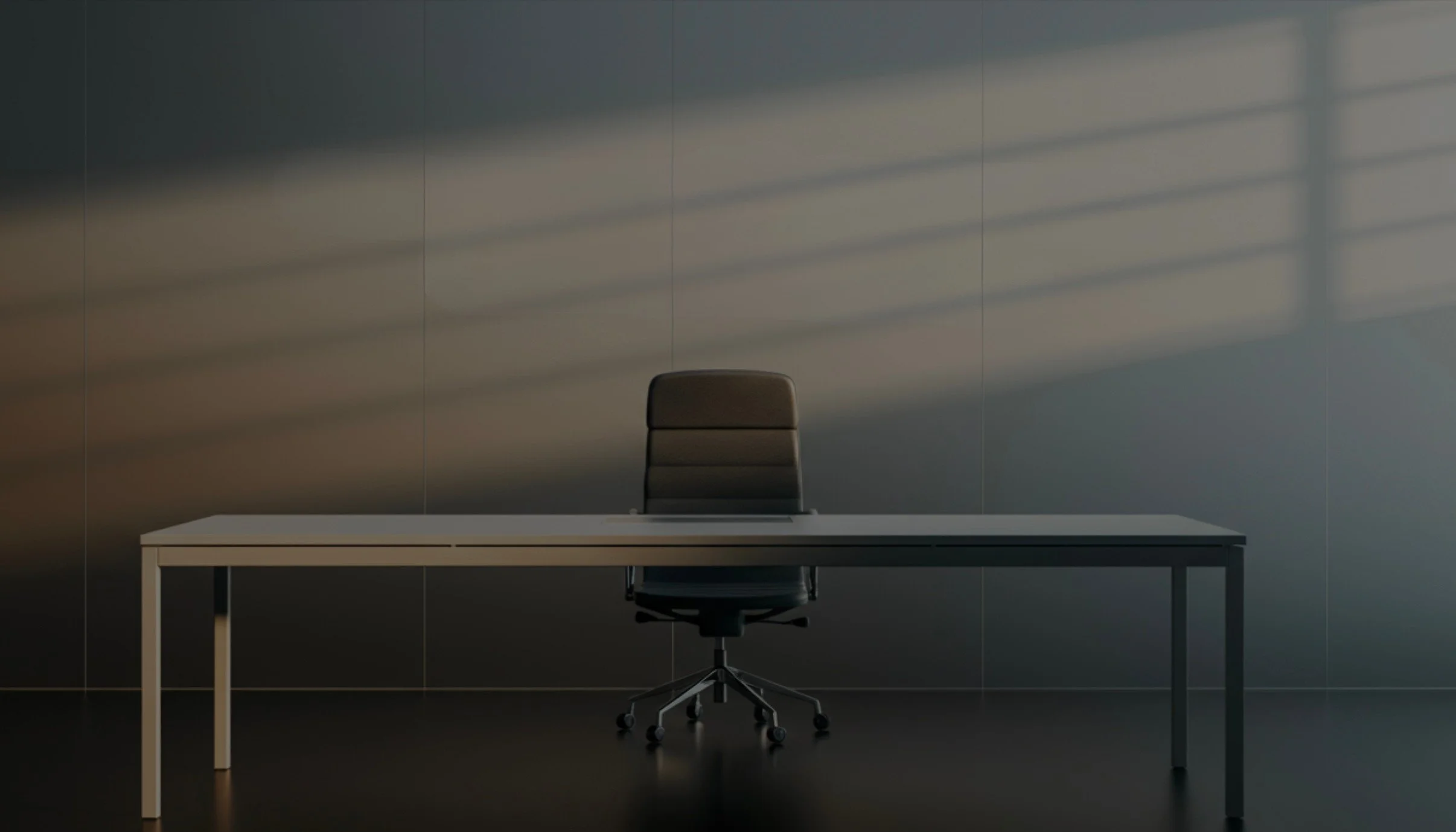 An empty office with a white desk, a black ergonomic chair, and light patterns from blinds on the wall.