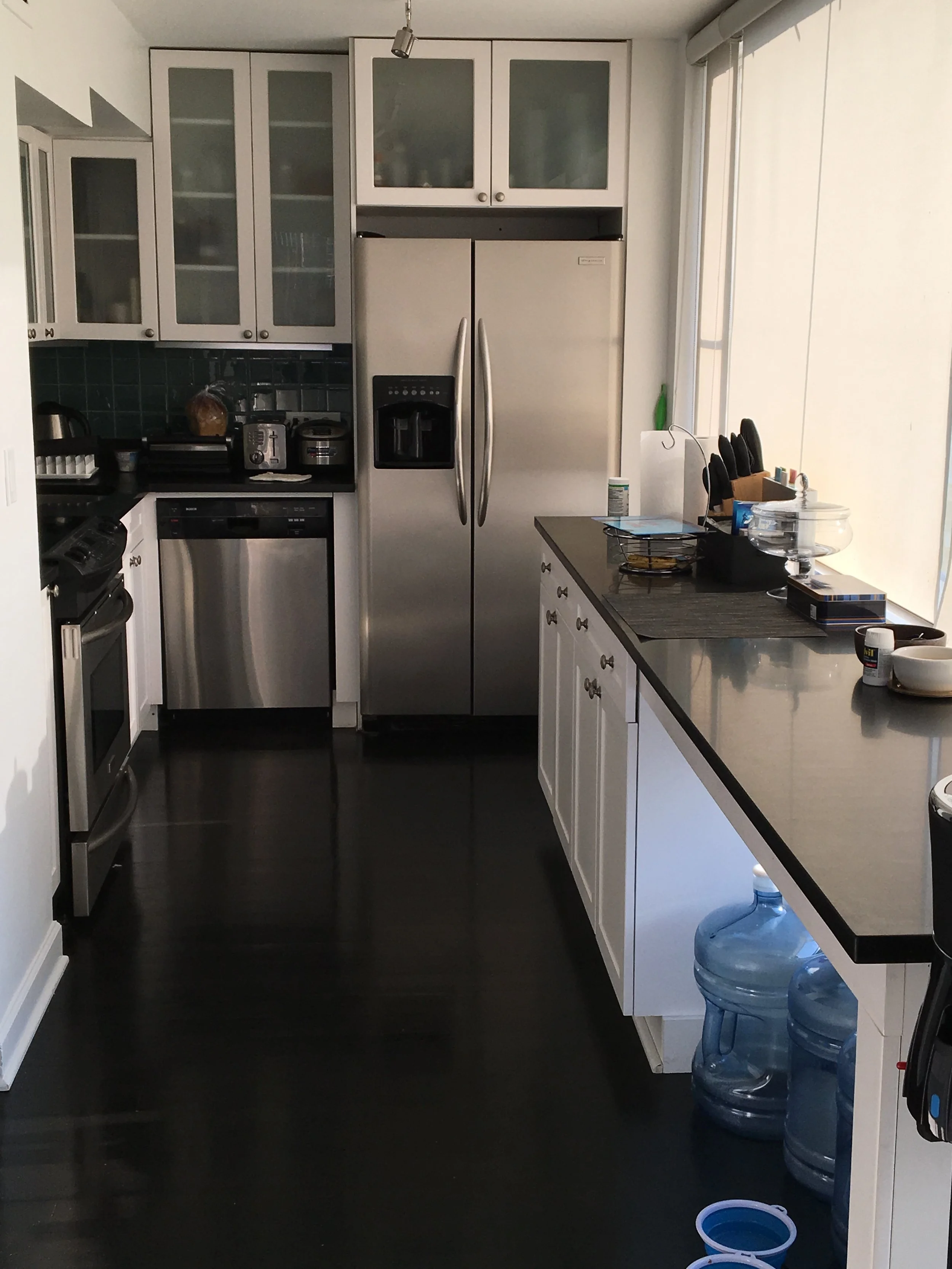 Modern kitchen with white cabinets and black countertops, stainless steel appliances including a refrigerator, dishwasher, and oven, black tiled backsplash, water jugs on the floor, and various small appliances and items on the countertops.