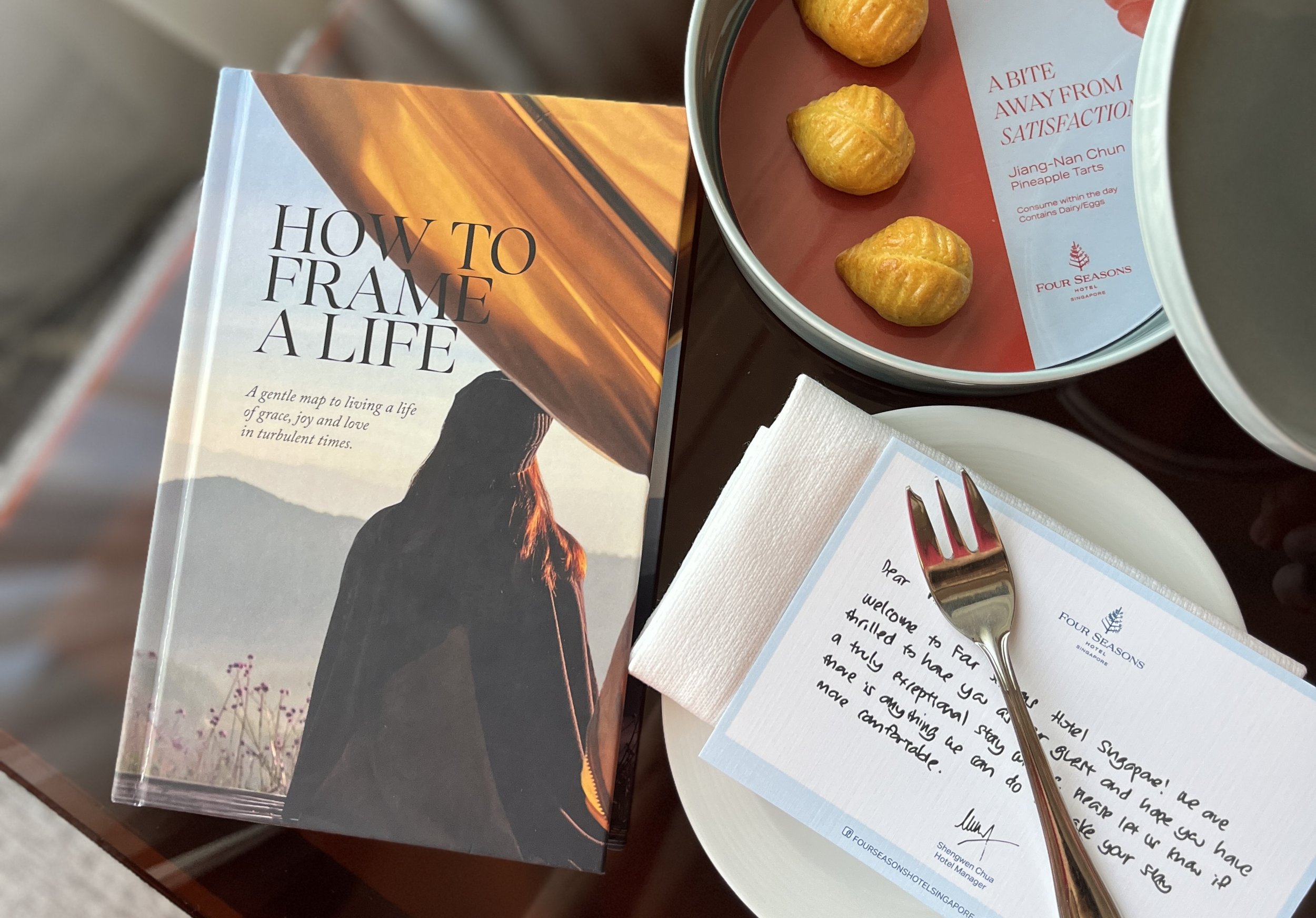 A book titled 'How to Frame a Life' is on a table next to a note card with a handwritten message, a fork, and a plate with four pineapple tarts, on a tray with a paper banner from Four Seasons Hotel Singapore.