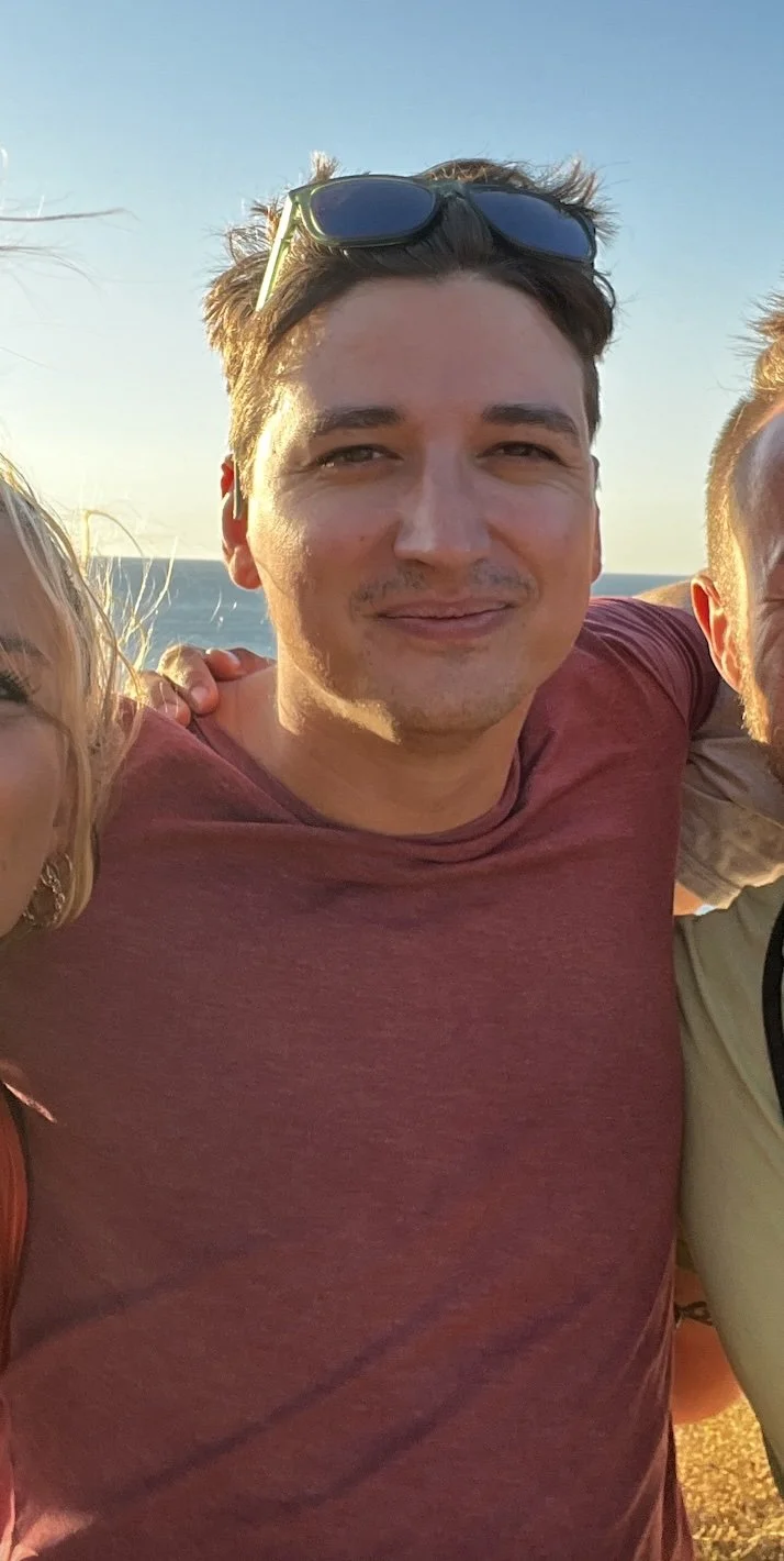 A young man with sunglasses on his head, smiling, wearing a red T-shirt, standing outdoors during sunset with the ocean in the background.