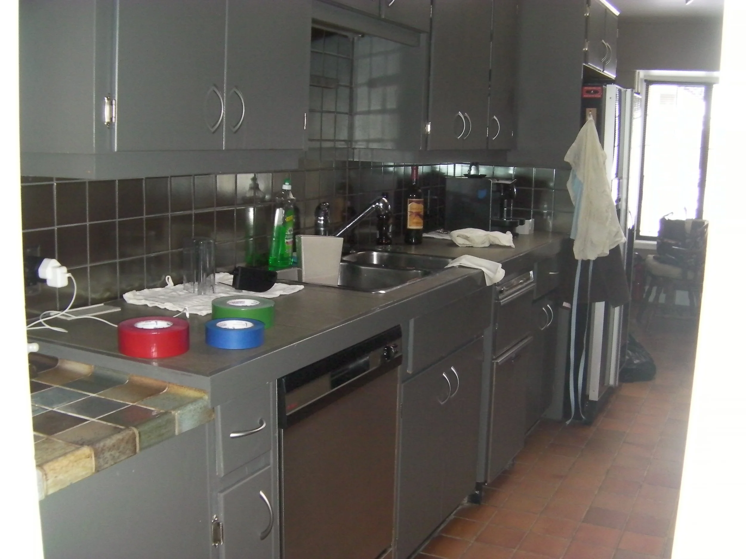 A cluttered kitchen with gray cabinets, a black tiled backsplash, and a stainless steel countertop. Items on the counter include green dish soap, cups, a white cloth, a bottle of alcohol, and a toaster. There are three rolls of tape in red, blue, and green on the counter. A dish towel hangs from a cabinet handle, and there are chairs and various objects near a window in the background.