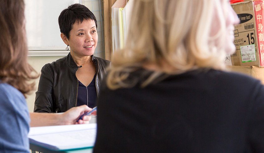 Three women in a discussion, one woman with short black hair facing the camera and others with blurred faces, in a room with shelves and boxes.
