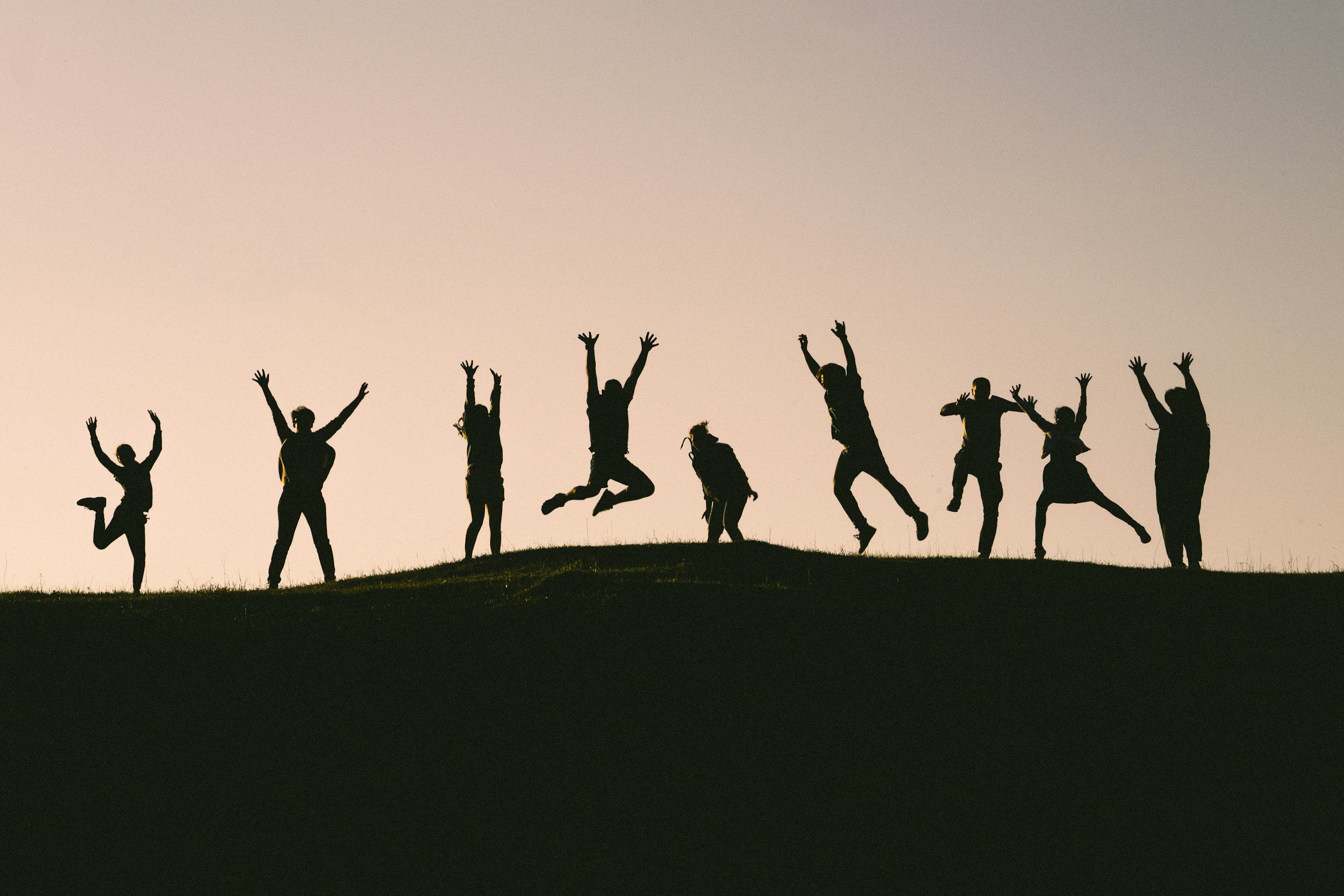 Silhouettes of nine people jumping and raising their hands on a grassy hill at sunset or sunrise.