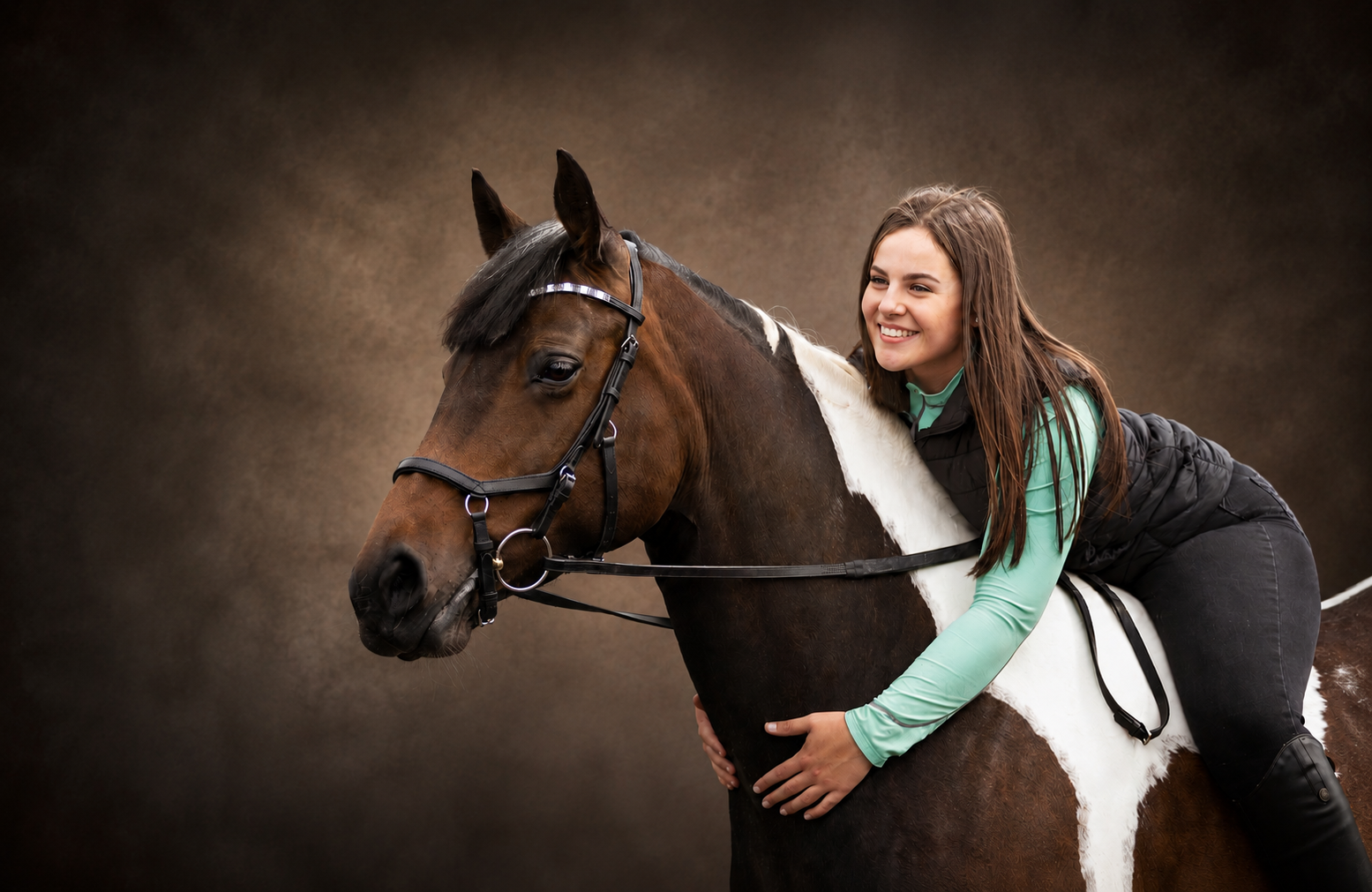 A young woman with long brown hair smiling while riding a brown and white horse, with a dark neutral background.