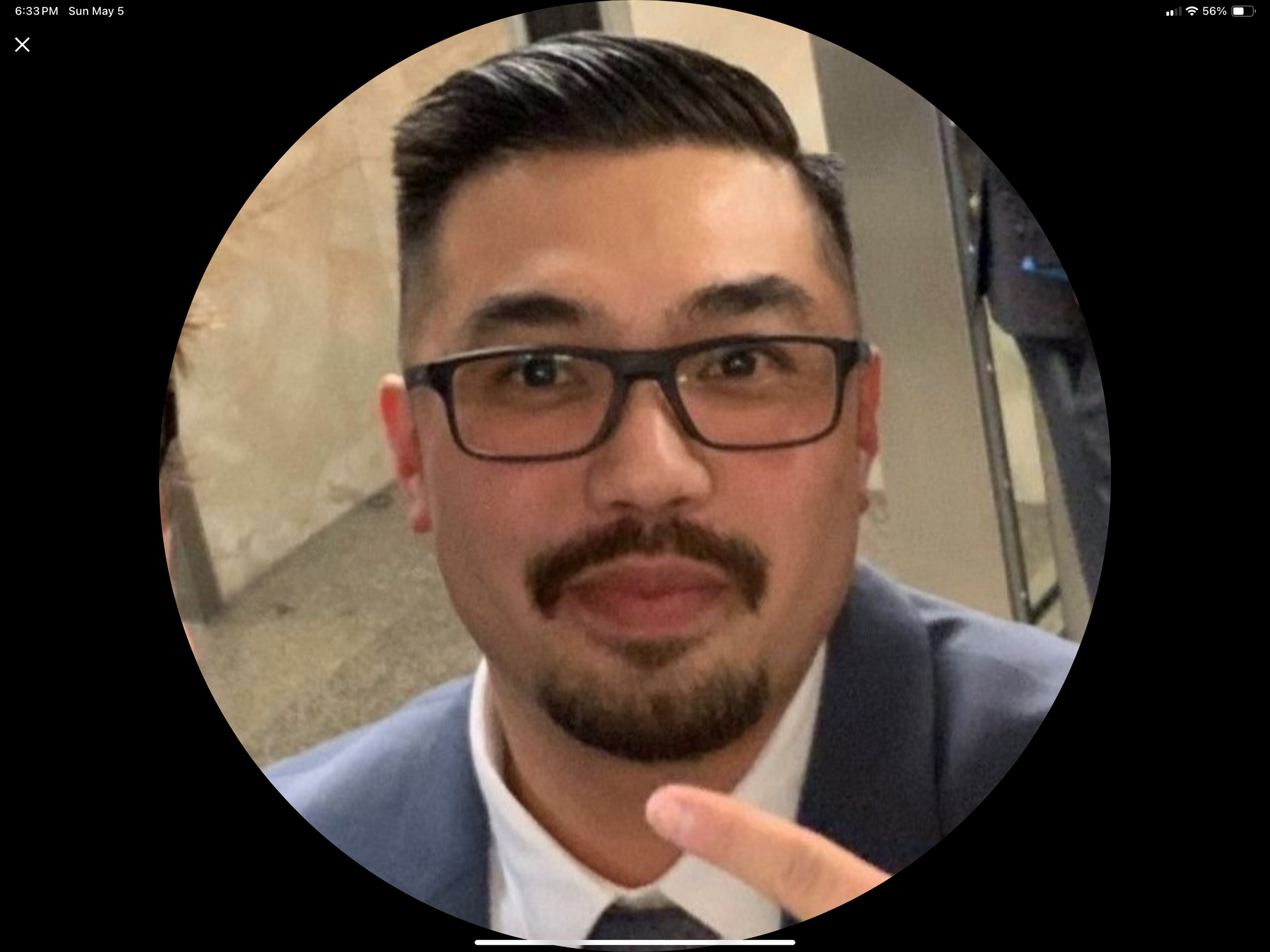 A man with black hair, glasses, and a beard and mustache, dressed in a suit, looking at the camera in an indoor setting.