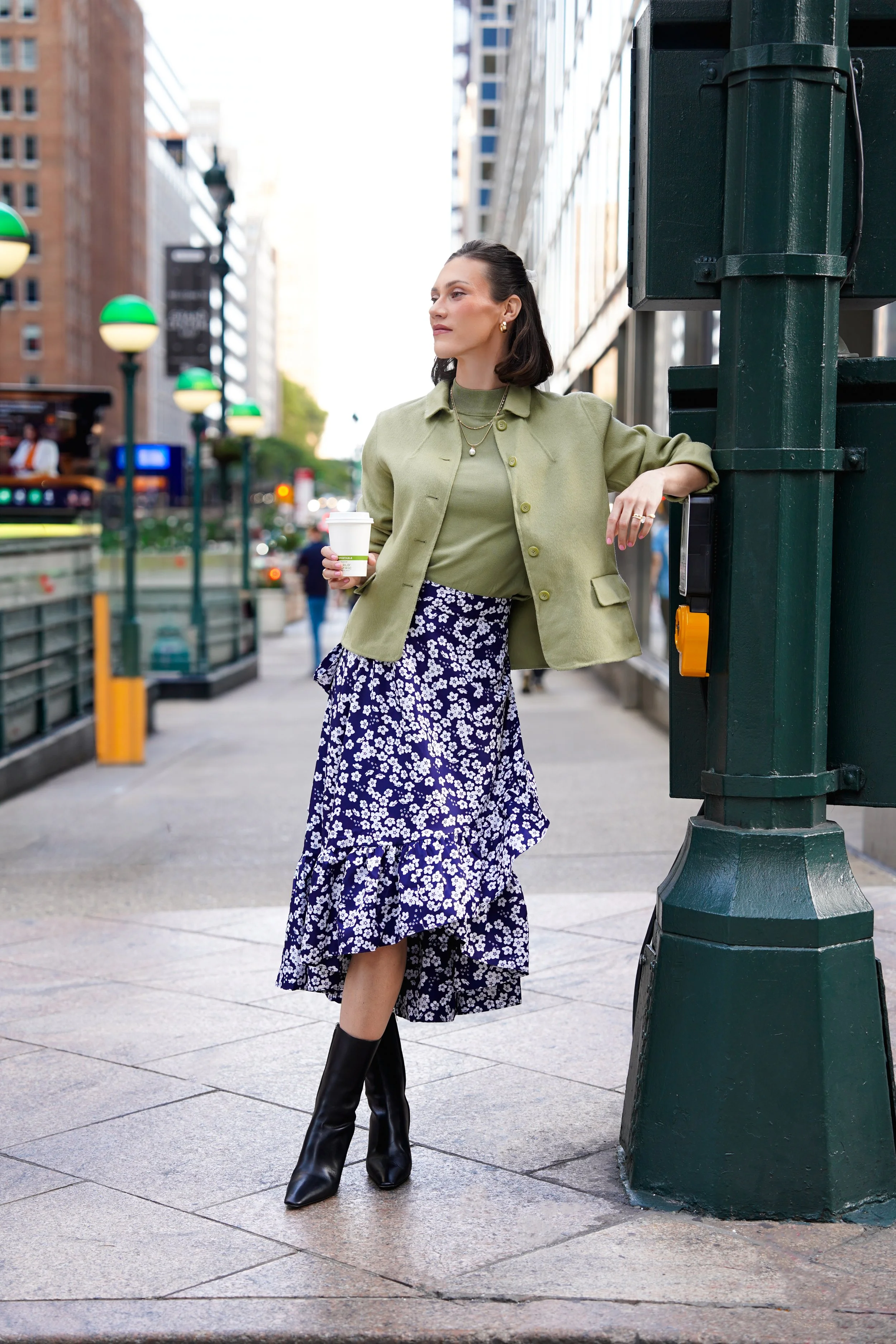 A woman with dark hair, wearing a green jacket, green top, floral skirt, and black boots, leaning against a green utility pole on a city sidewalk holding a coffee cup.