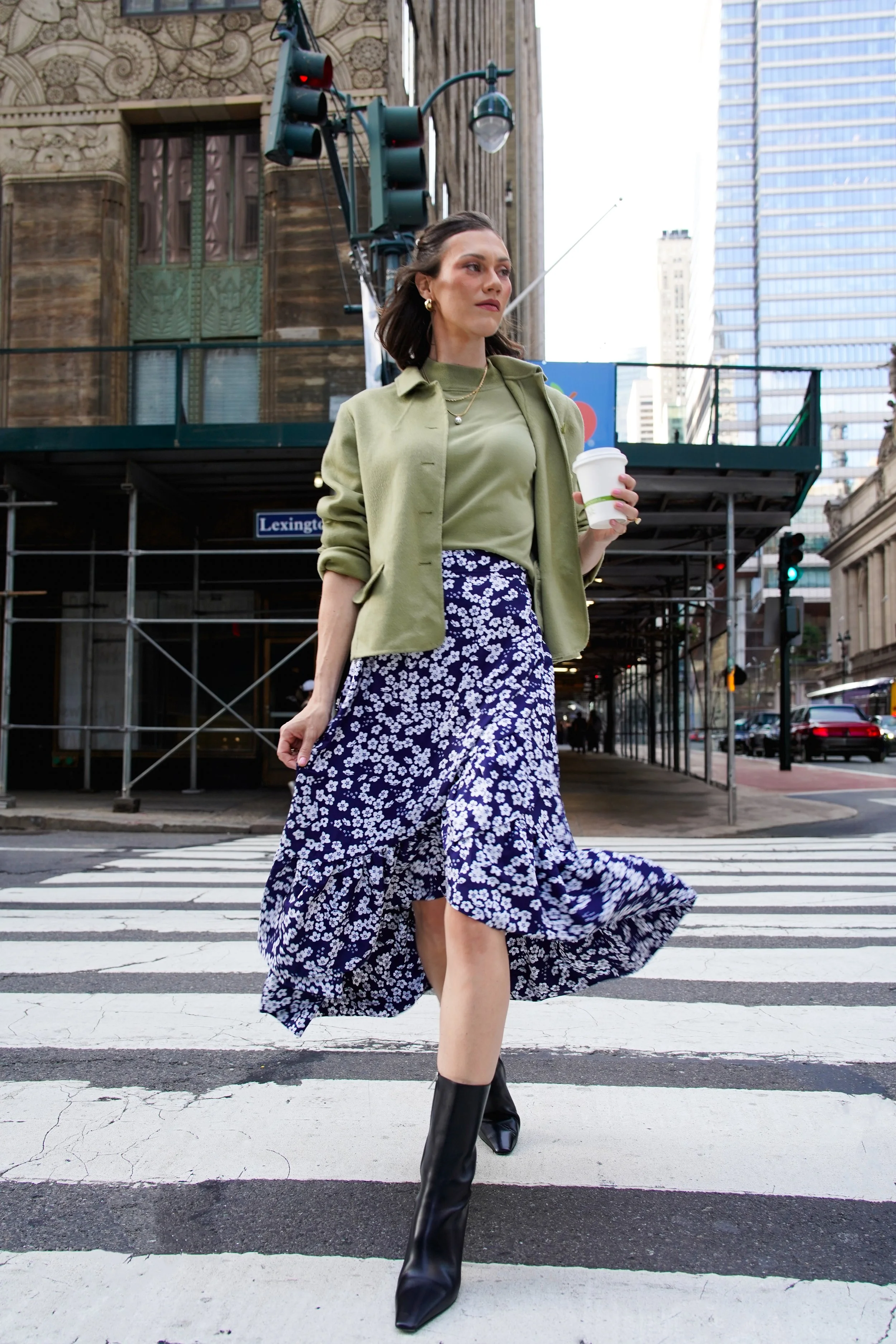 A woman in a green jacket and floral maxi skirt crossing a city street with tall modern buildings and a stoplight in the background, holding a coffee cup.