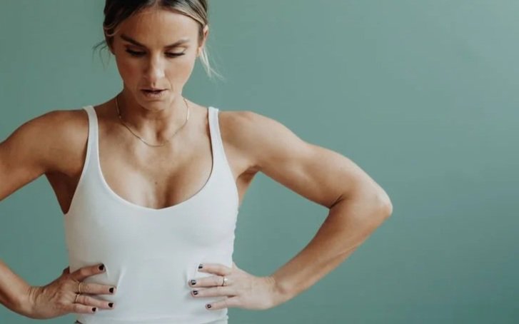 Woman with blonde hair in a white tank top looking down with hands on her hips against a green wall.
