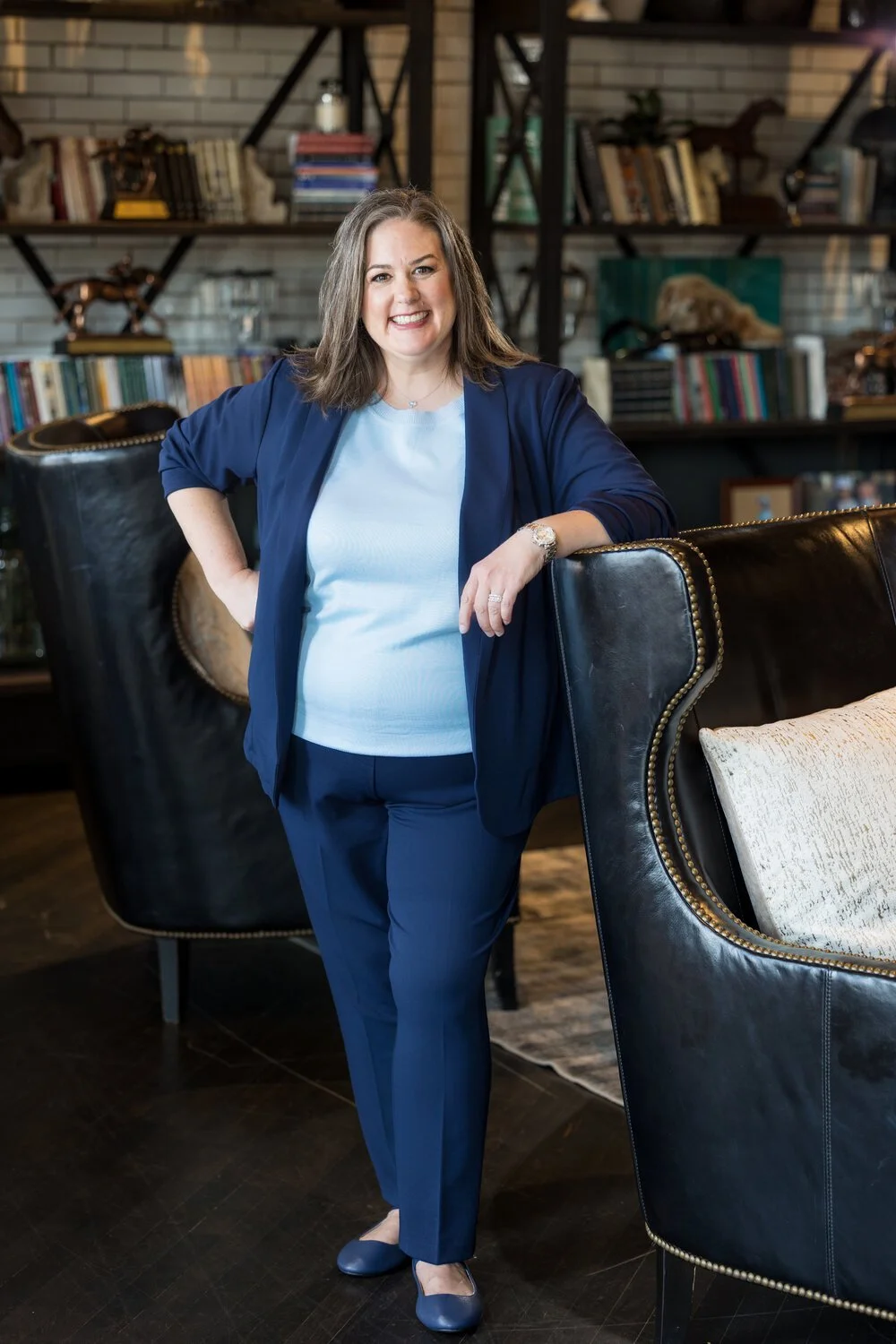 Amy Simpson with shoulder-length brown hair, smiling, wears a light blue top, dark blue blazer, and matching pants, leaning against a black leather chair in a cozy room with bookshelves behind her.