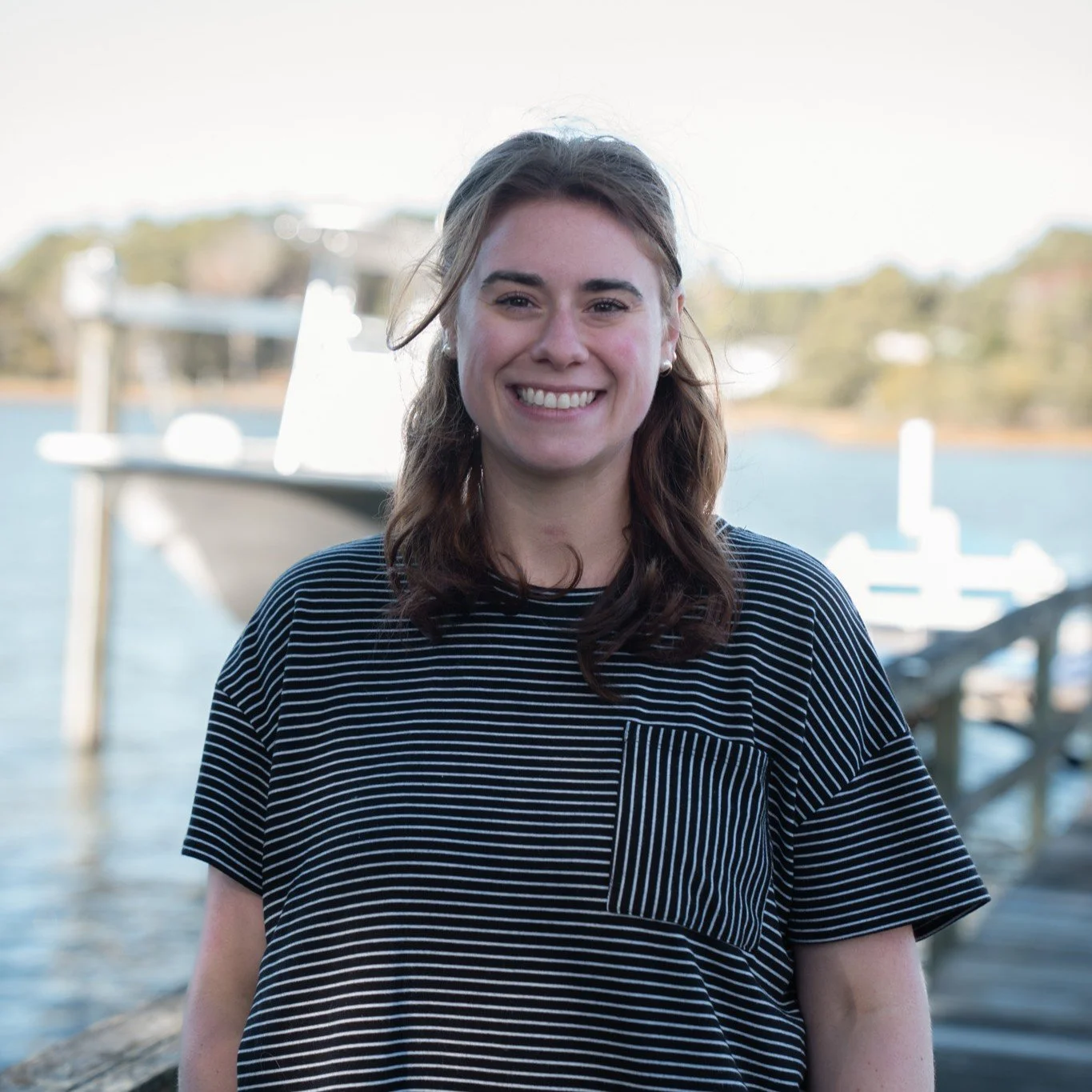 Jade Westmoreland smiling wearing a black and white striped shirt. She is standing on a pier with a boat and water in the background.