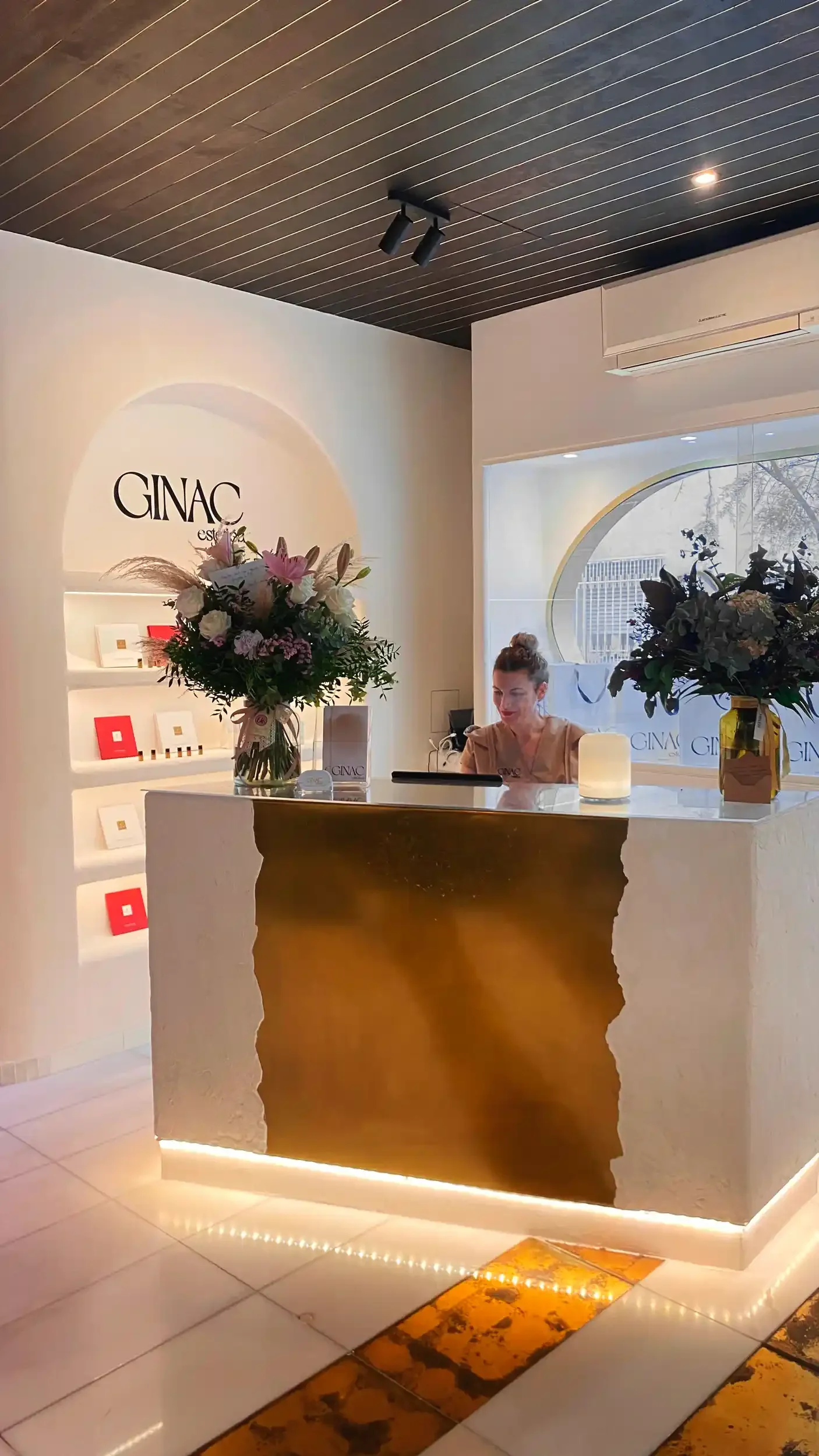 Mostrador de la recepción de Ginac Estética con dos arreglos florales y una mujer atendiendo, paredes blancas, exhibiciones y una ventana redonda con vista al exterior, iluminación cálida.