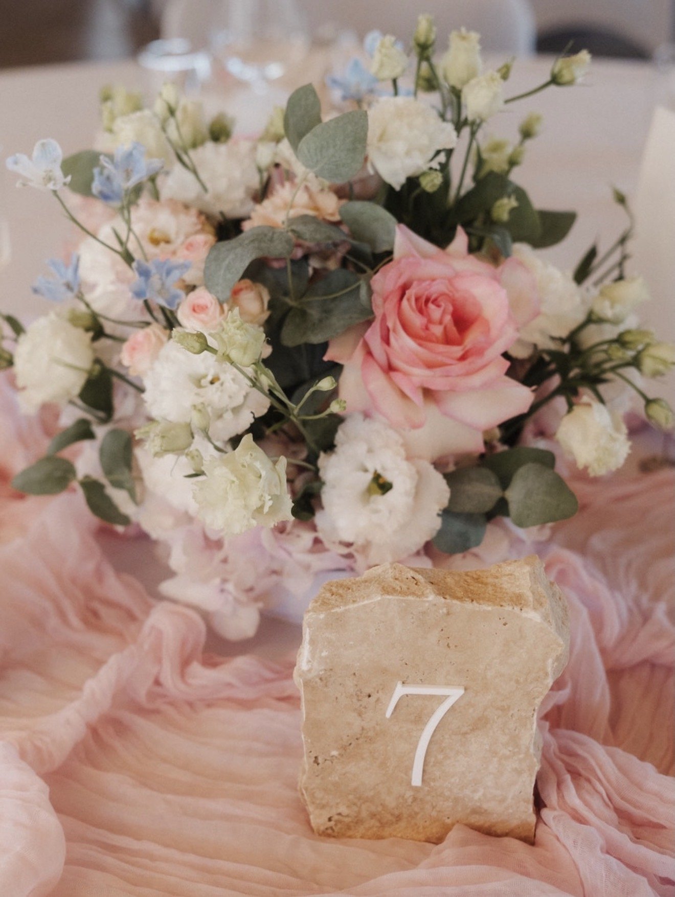 Blumenstrauß mit weißen, rosafarbenen und blauen Blumen, Eukalyptusblättern, auf rosa Tuch, mit Steinschild mit der Nummer 7 im Vordergrund.