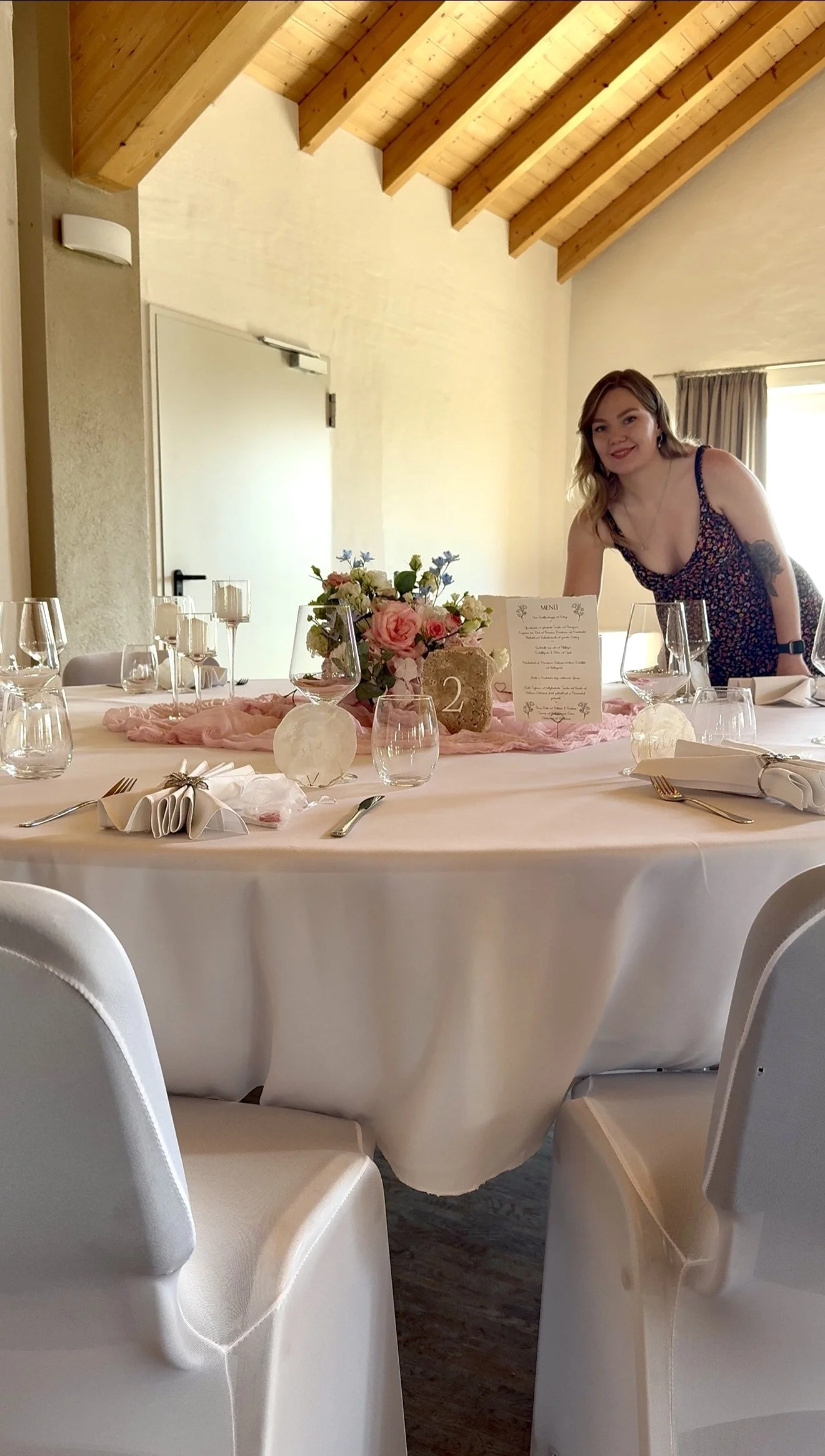 Ein festlich gedeckter Esstisch mit Blumen, Kerzen und einem Menü für eine Feier, im Hintergrund eine junge Frau mit einem schwarzen Kleid mit Blumenmuster, die an einem festlich gedeckten Tisch steht.